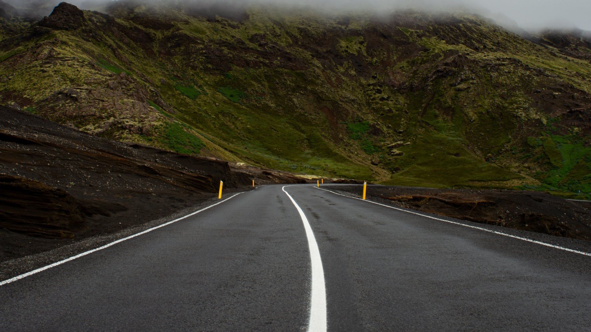 Road leading into a mountainous landscape; hills are covered with green vegetation, with low-lying clouds.