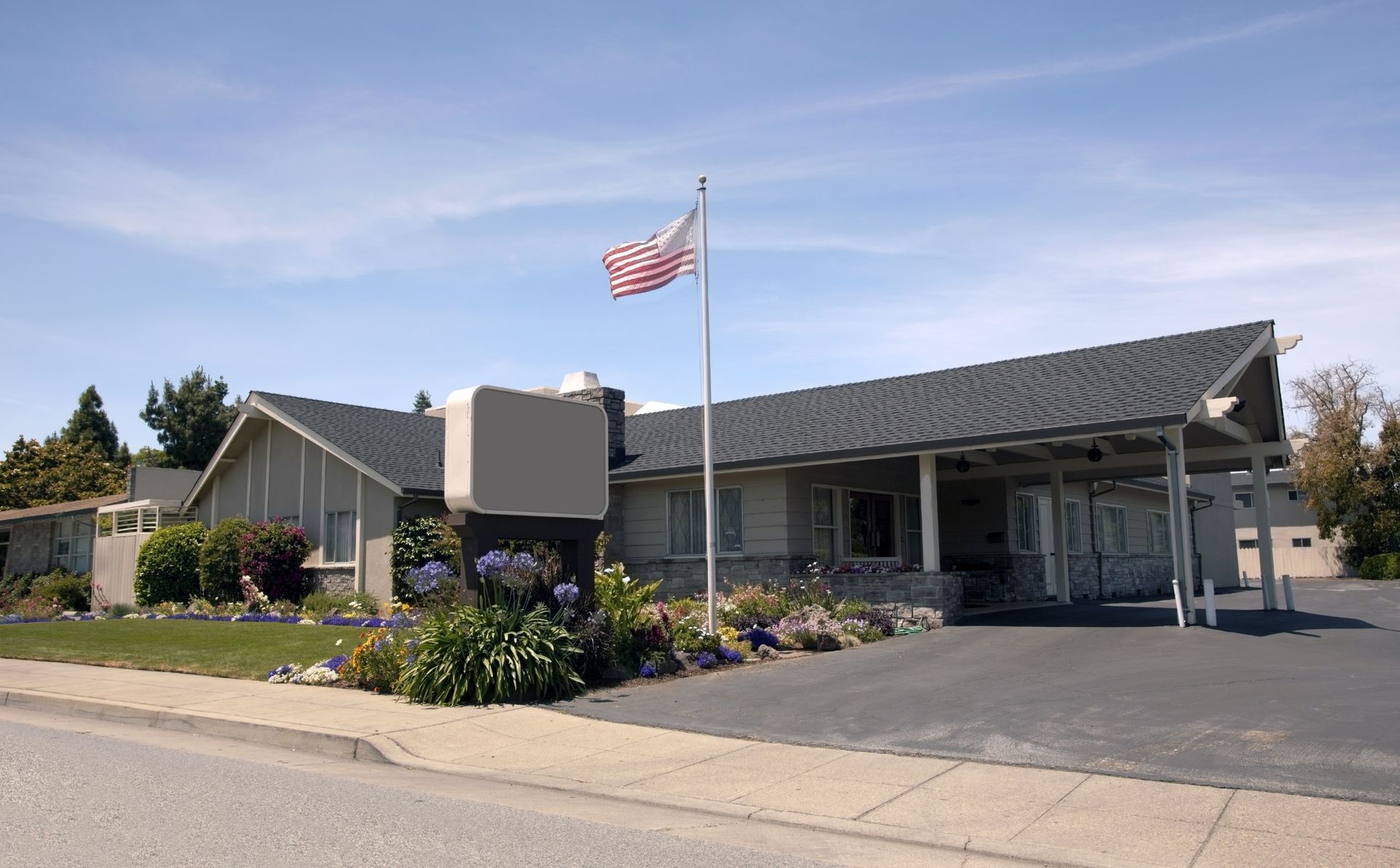 A one-story building with a carport and American flag. Dark roof, gray walls, and a driveway.