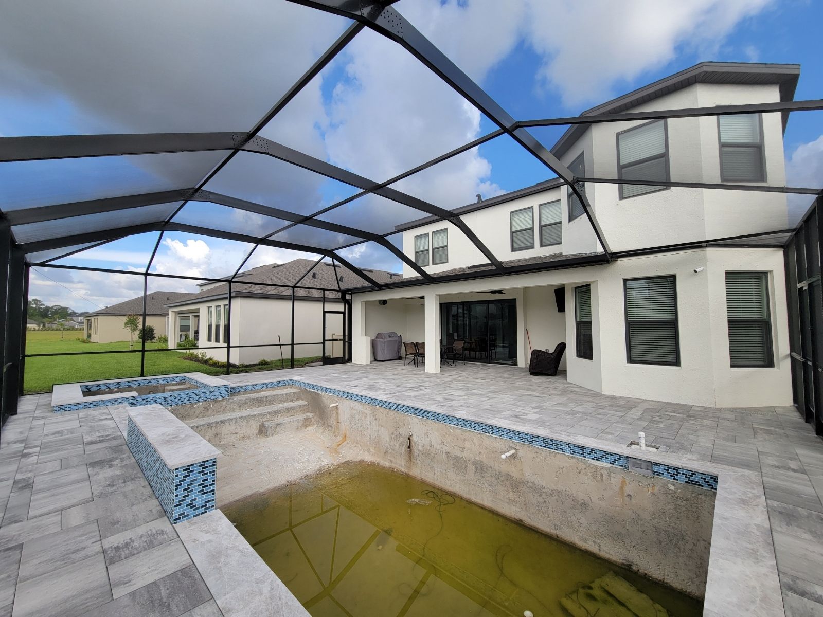 Covered patio with a rectangular pool beside a white house under a screened enclosure.