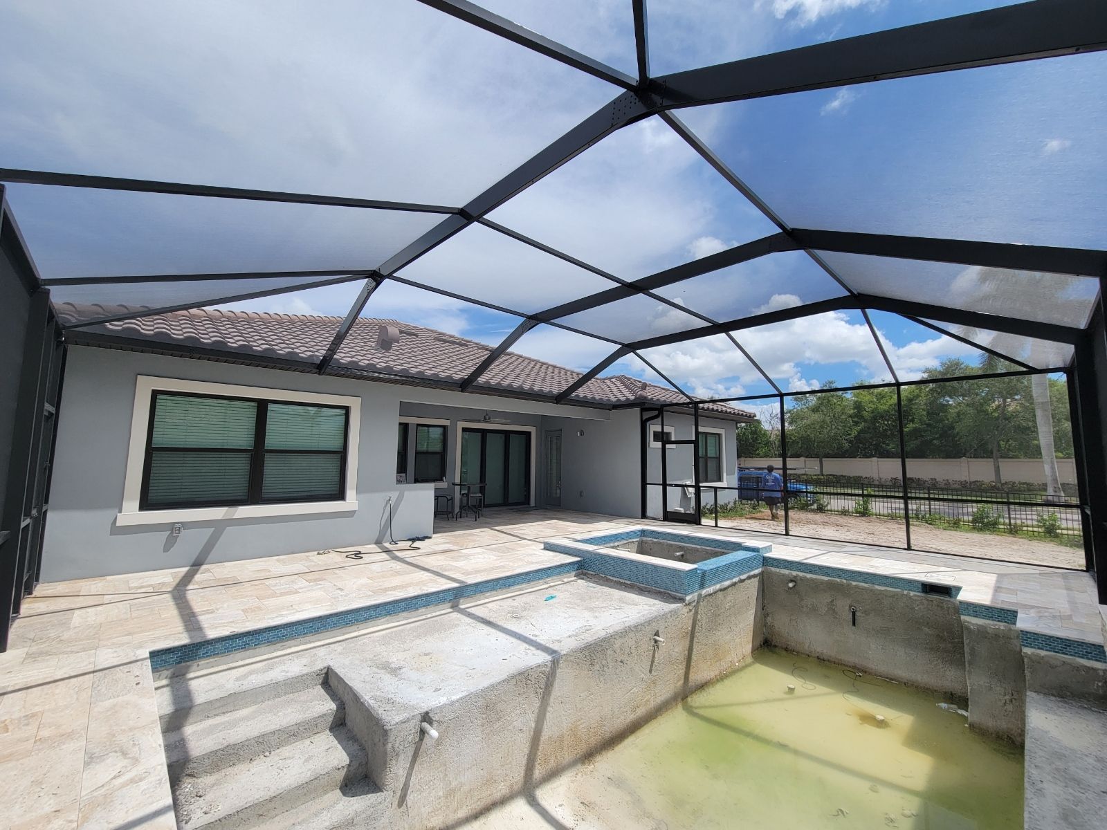 Screened pool patio with concrete deck, steps, windows, and a partially filled pool under blue sky.