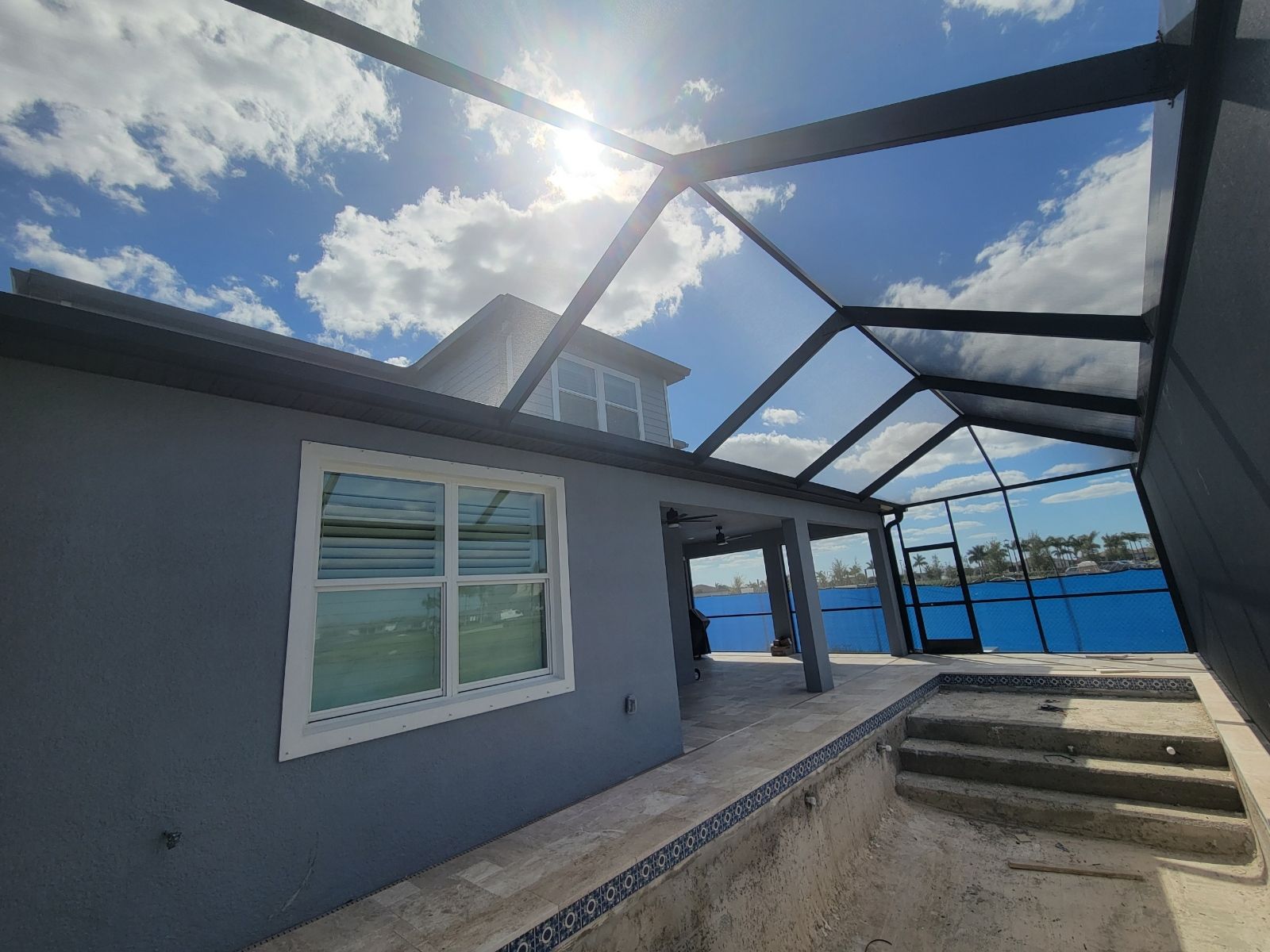 Back patio with gray wall, white window, and blue pool under a sunlit metal frame structure