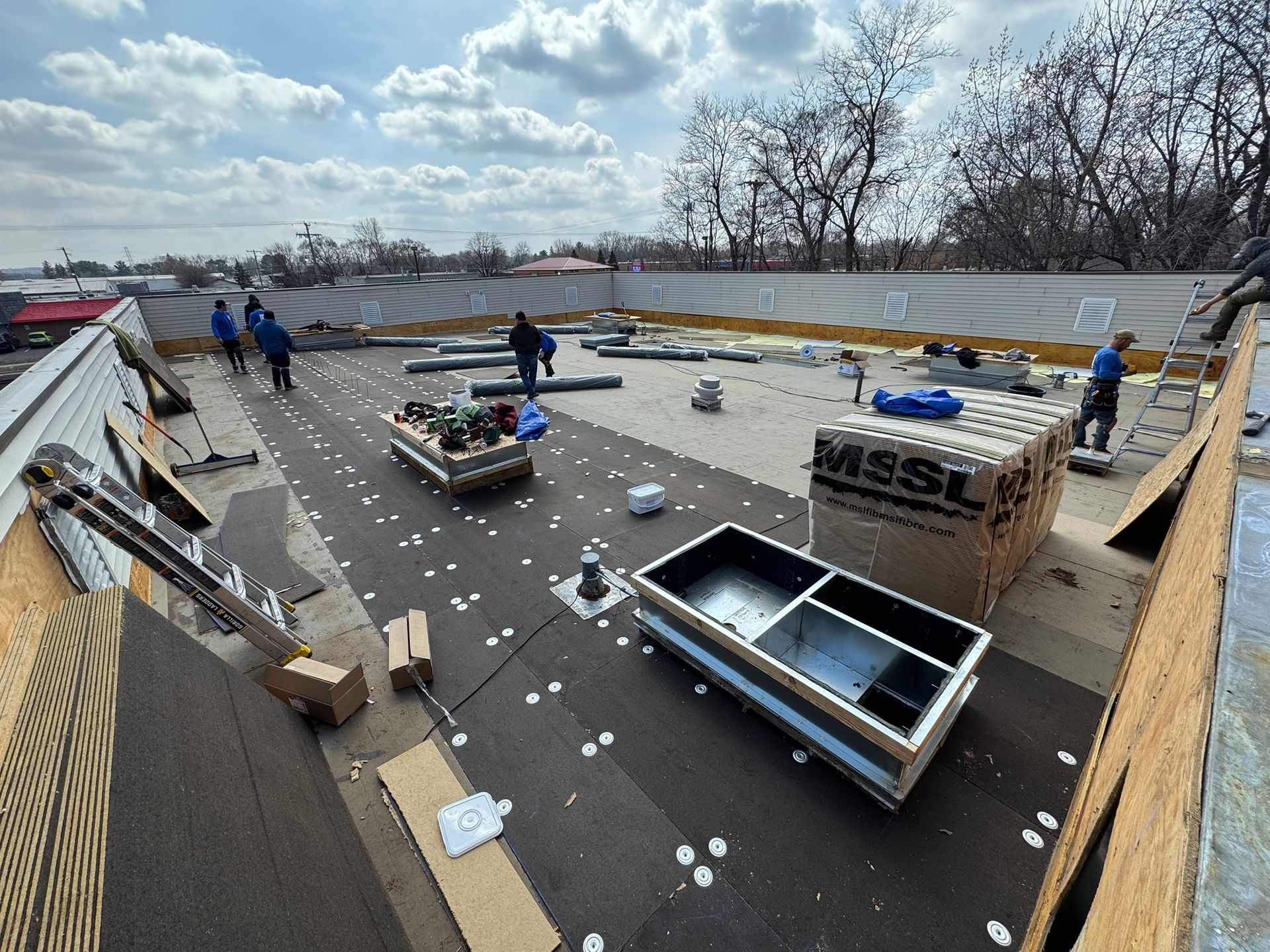 Roof under construction with workers, scattered materials, and open skylights under a cloudy sky