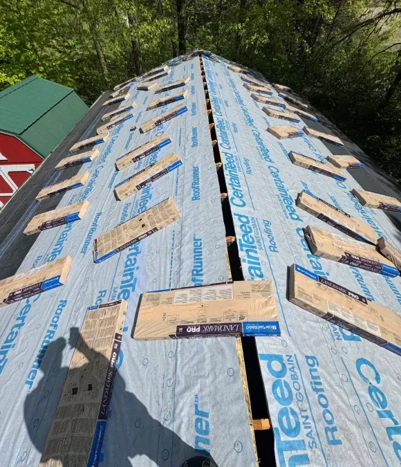 Roof under construction; blue underlayment and wooden battens. Sunlight and trees in background.