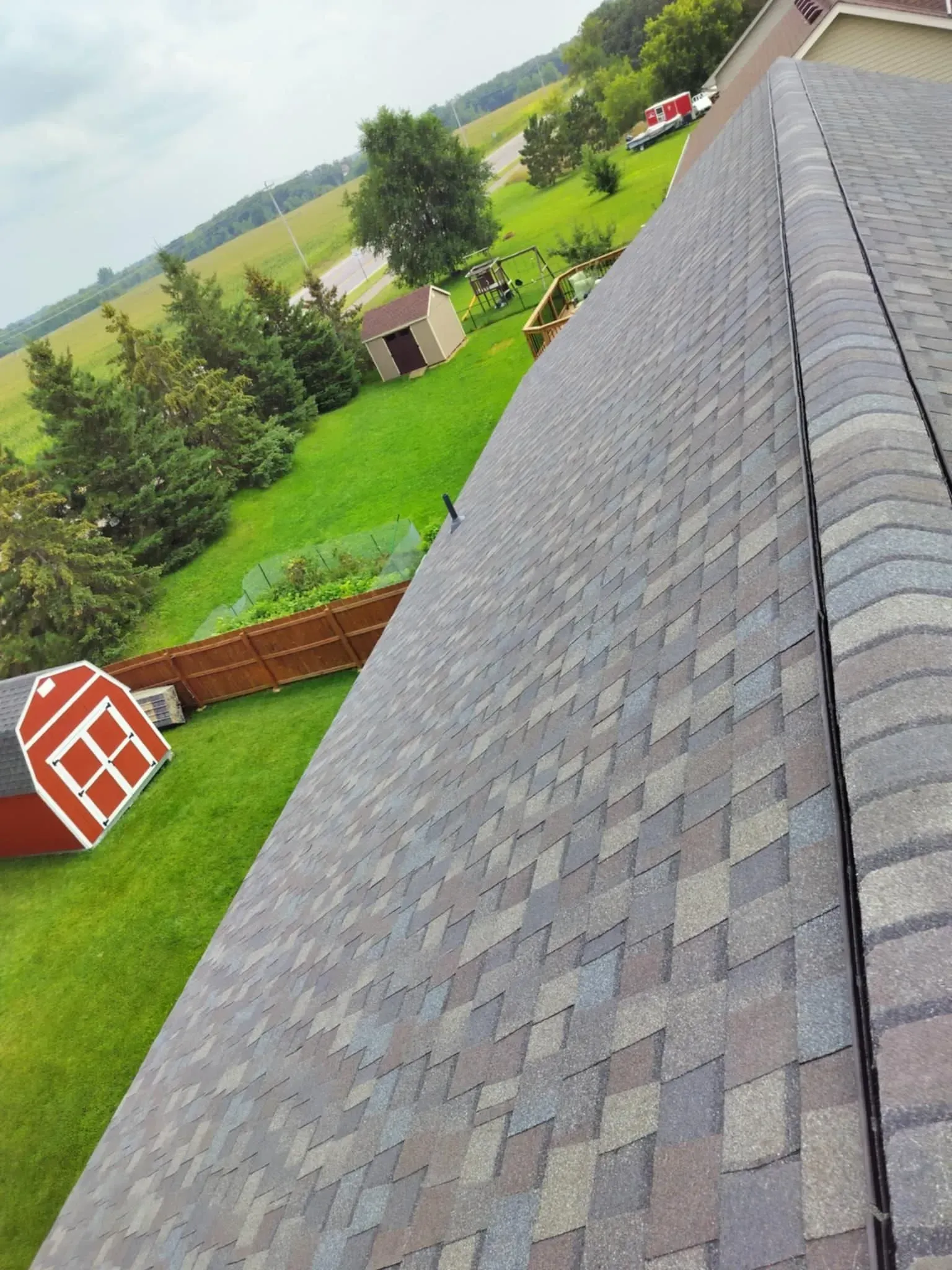Asphalt shingle roof, with a view of a grassy yard, trees, and small buildings under an overcast sky.