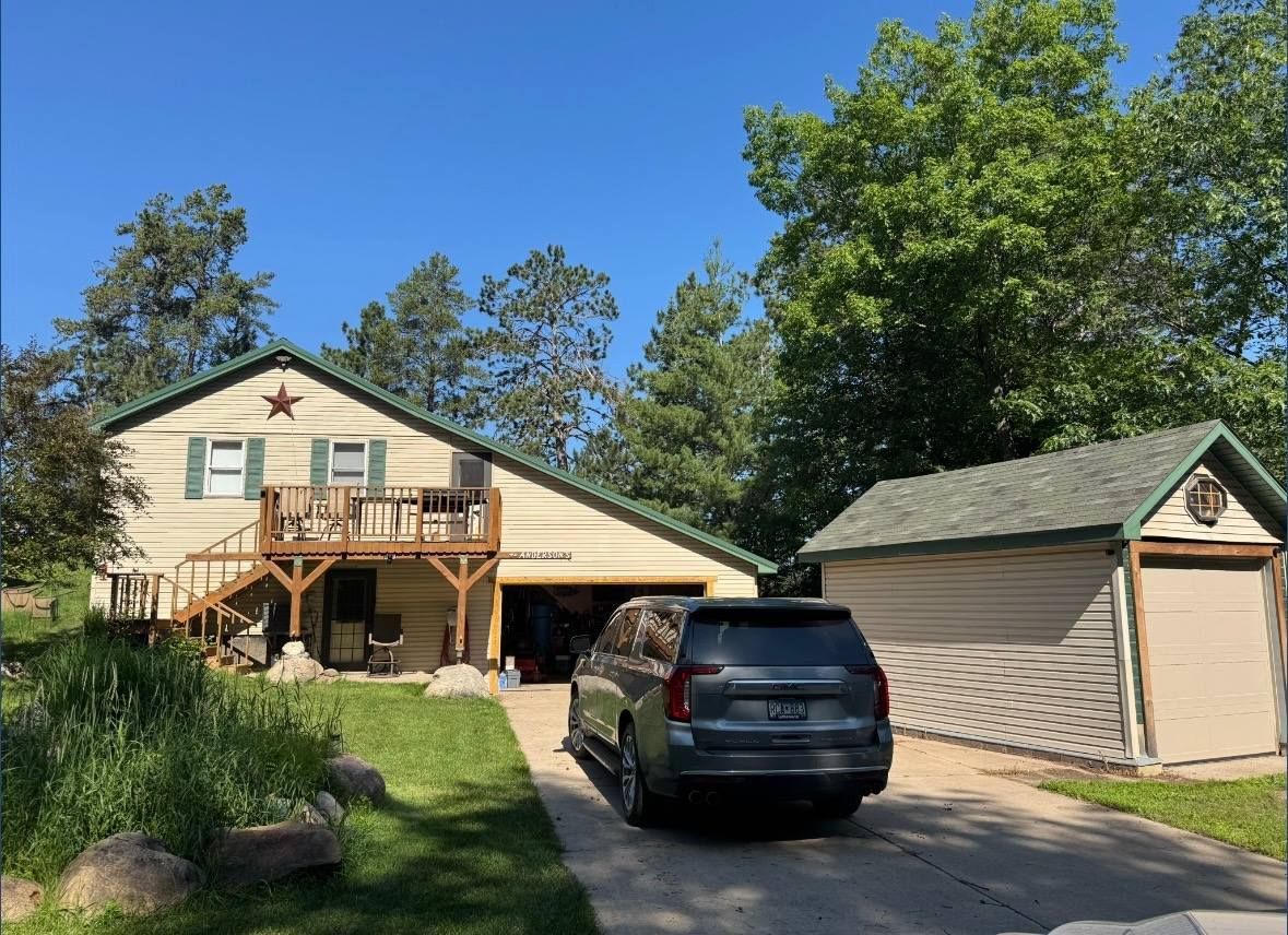 Tan house with attached garage, wooden deck, and parked SUV in driveway. Sunny day, green trees.