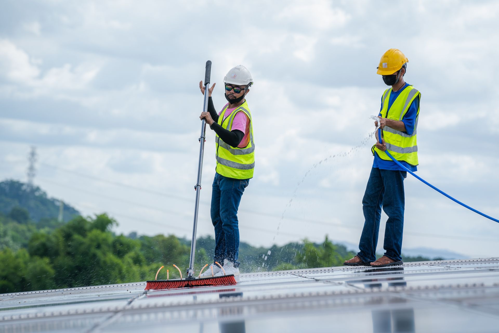 Two workers cleaning solar panels with a brush and hose on a rooftop, wearing safety vests and helmets.