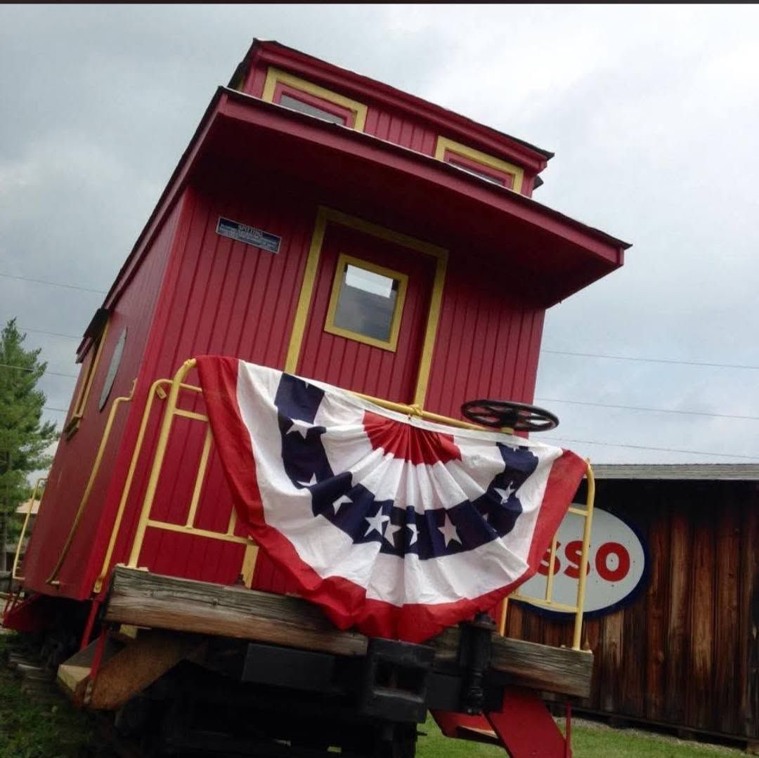 Tilted red caboose with patriotic bunting and yellow trim on a railroad track