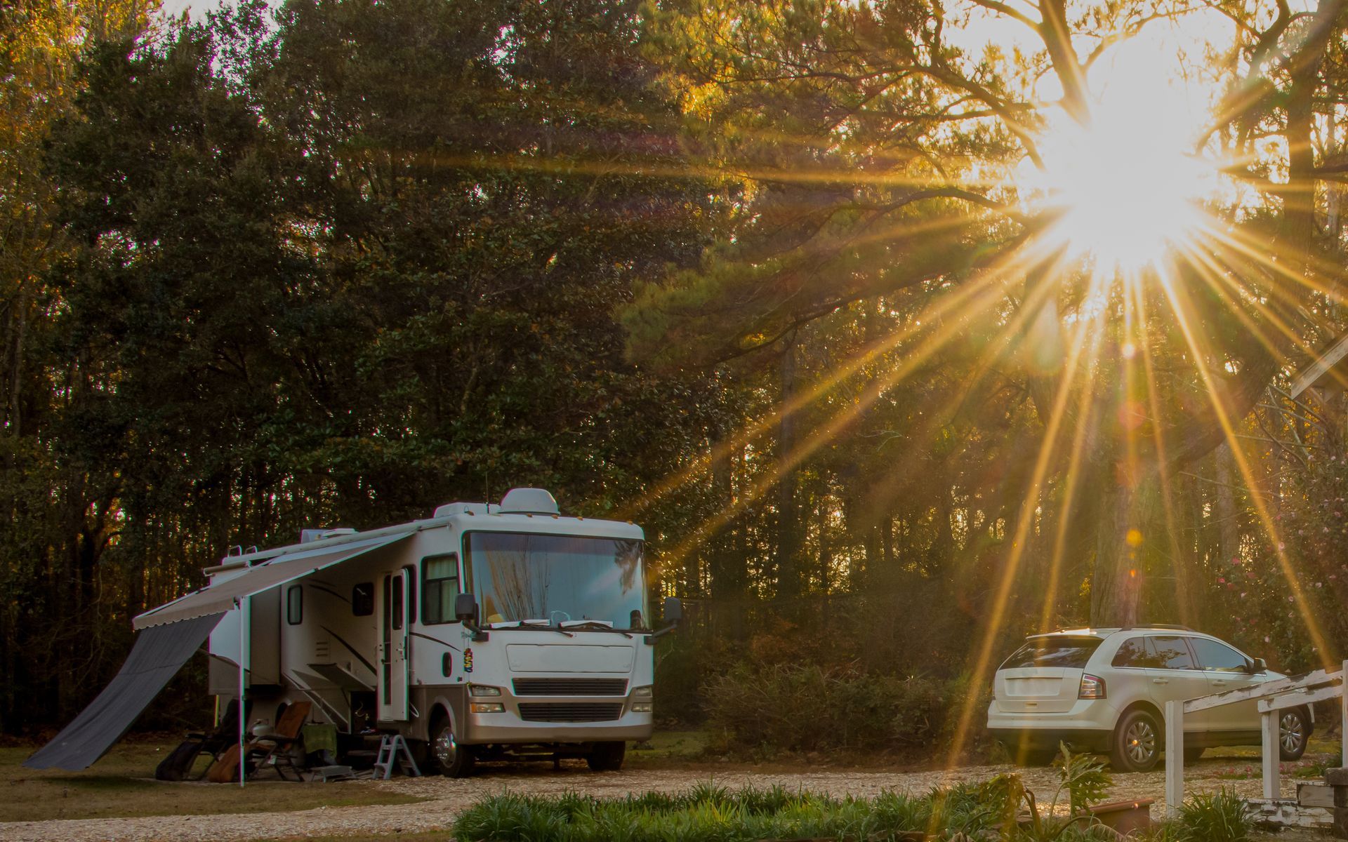 RV and white SUV parked at a wooded campsite under bright sunbeams at sunset.
