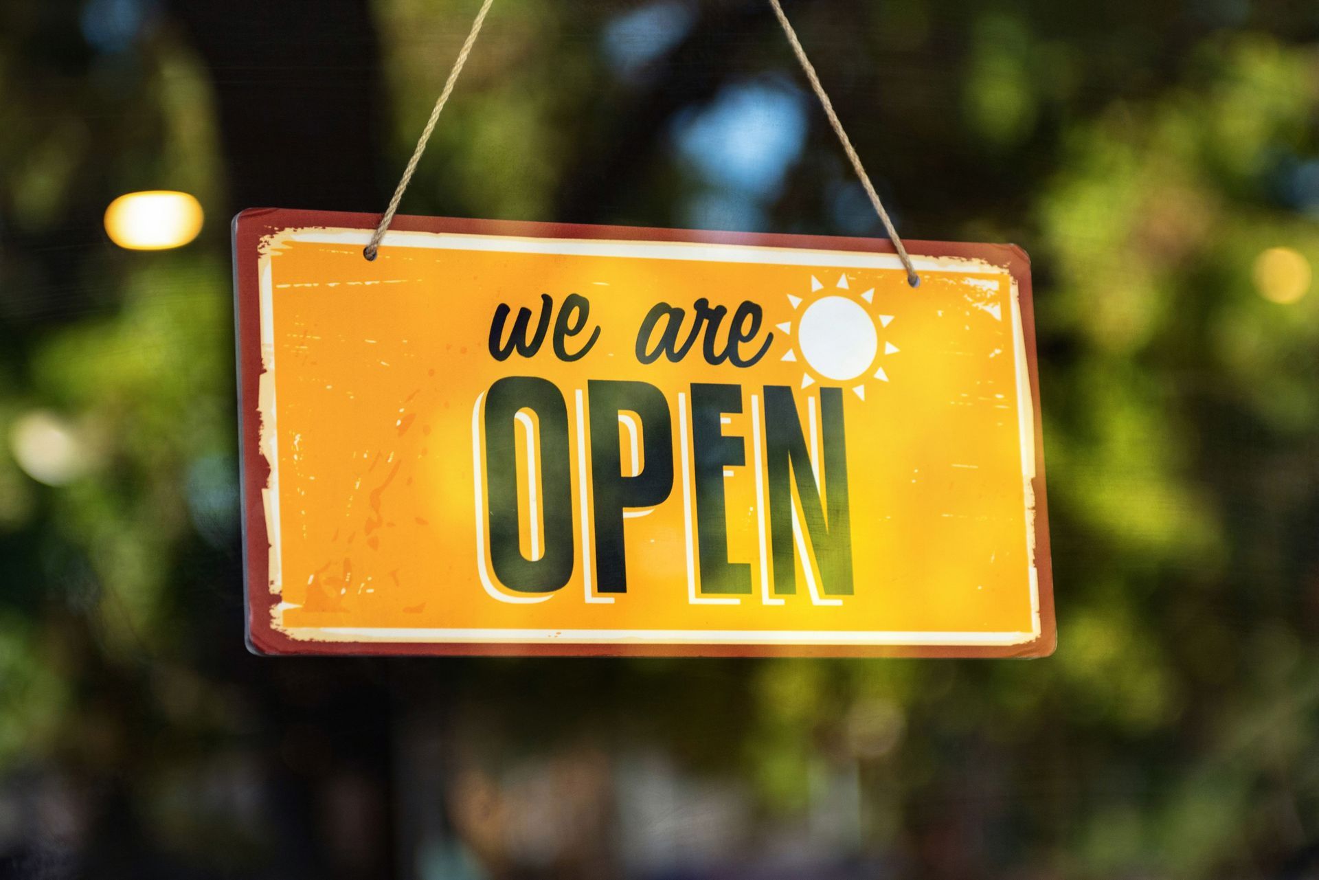 Orange hanging sign reading “we are OPEN” in black letters, with blurred greenery in the background