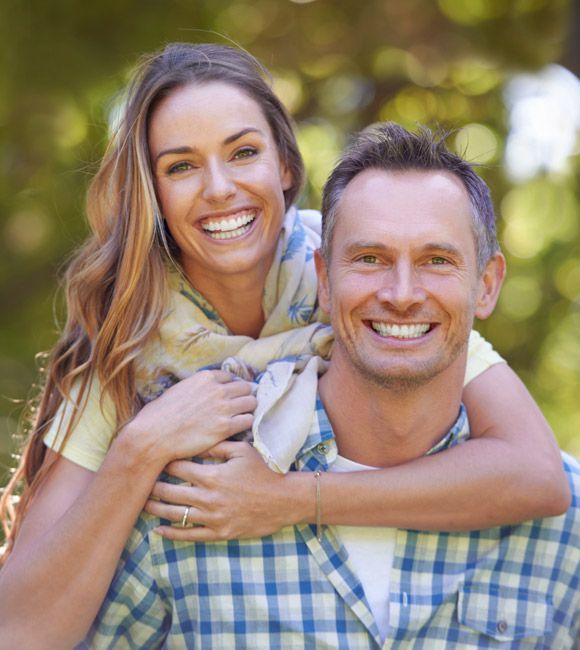 Smiling couple outdoors, the woman hugging the man from behind in a sunlit park.