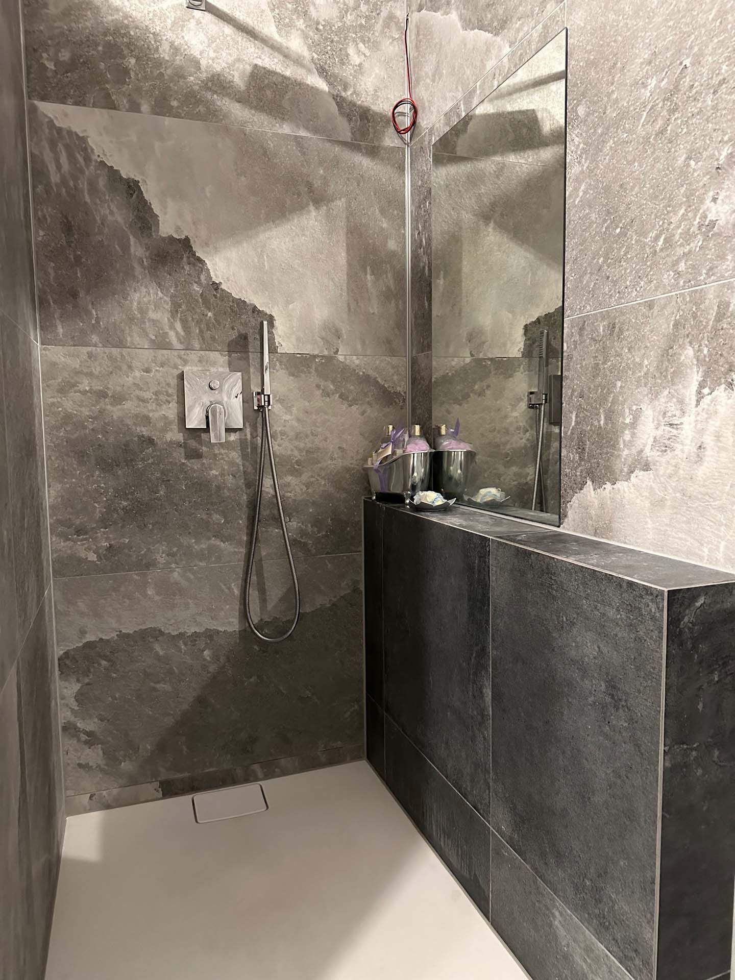 Ein Badezimmer mit edlen grauen Wandfliesen, ebenerdiger Dusche und großem Spiegel.