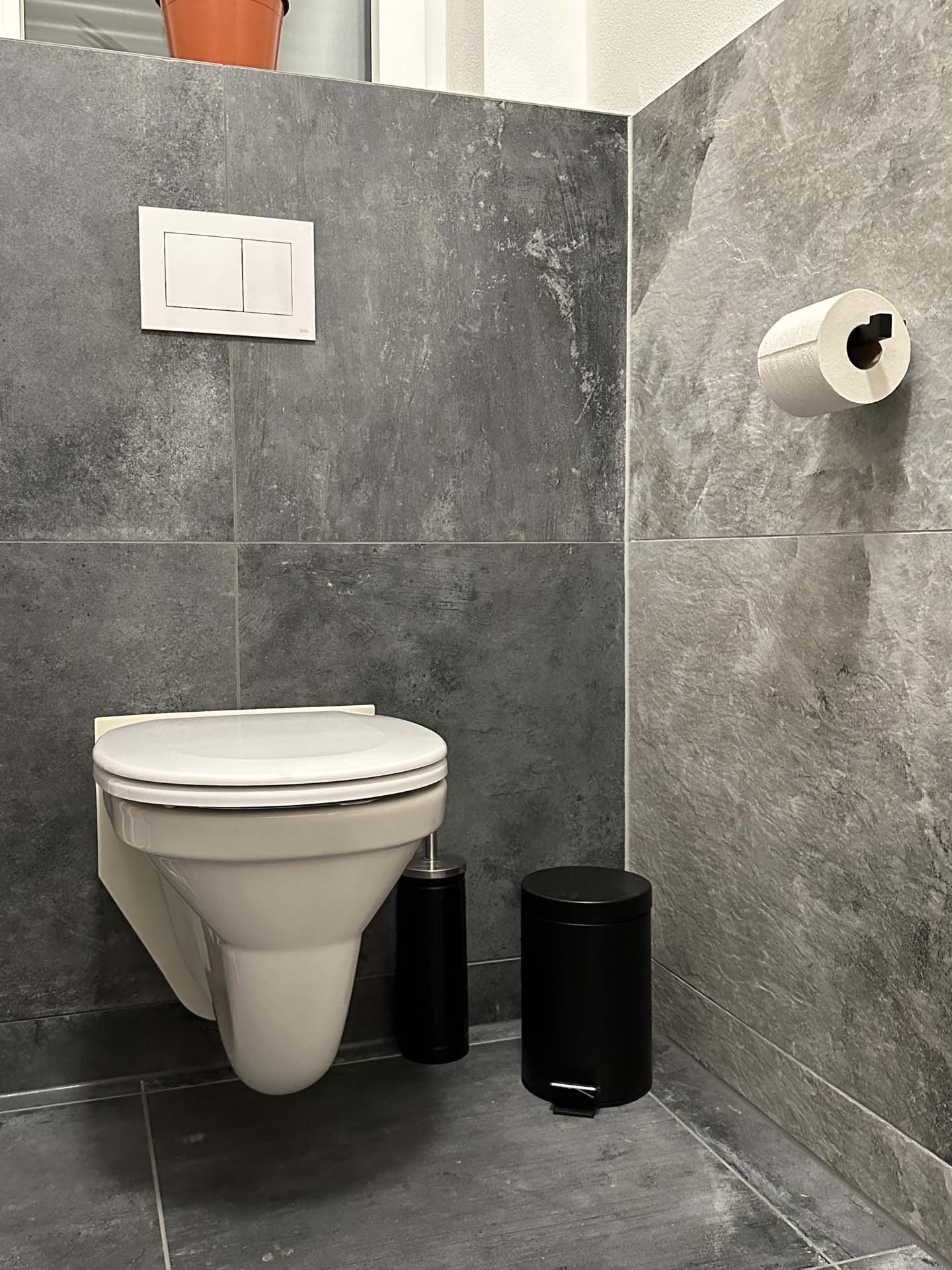 Ein Badezimmer mit grauen Wandfliesen, Toilette, Mülleimer und Toilettenpapier.