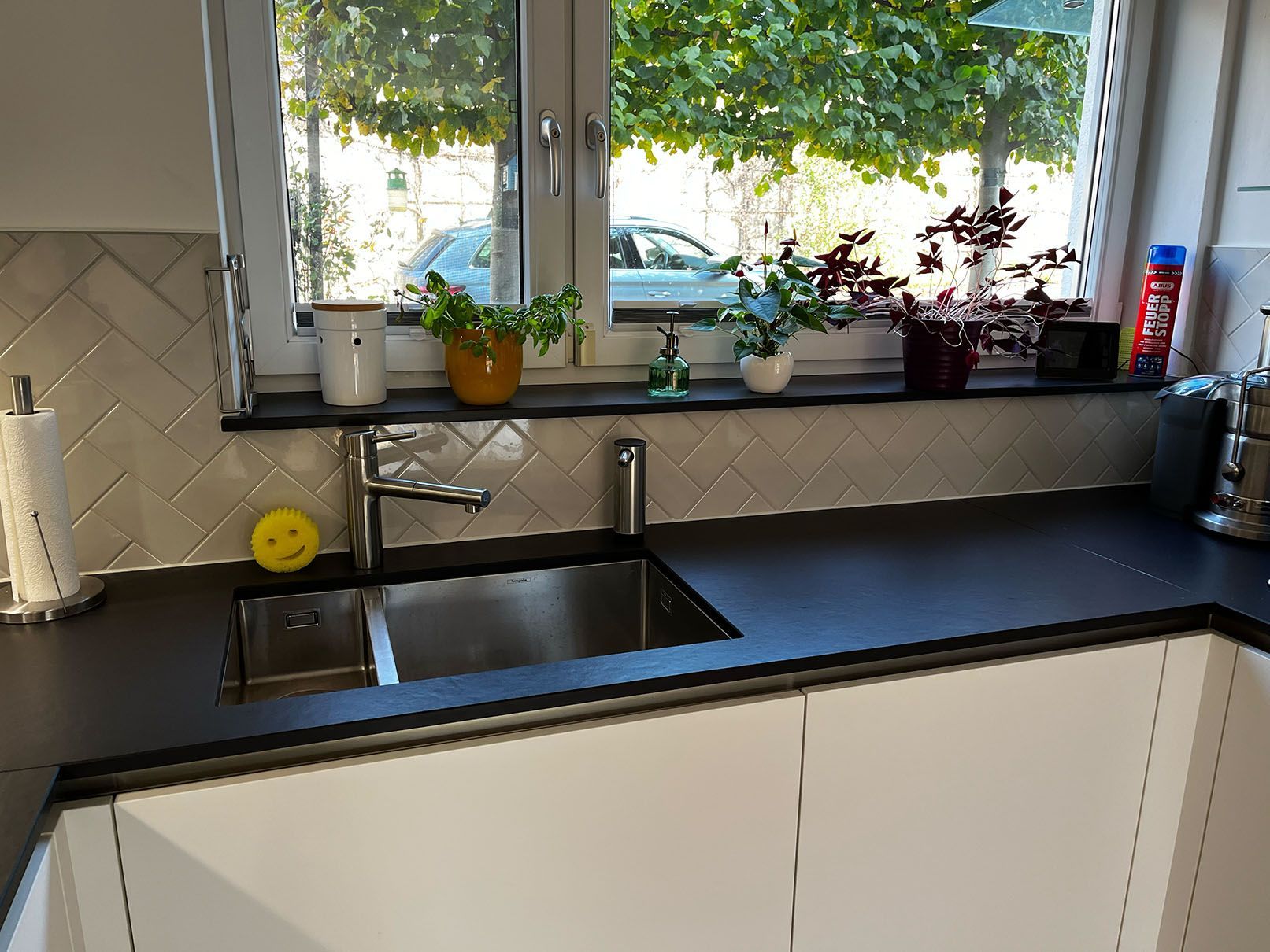 Eine Küche mit Spüle, mit Wandfliesen im Fischgrätenstil und ein Fenster mit Topfpflanzen auf der Fensterbank.
