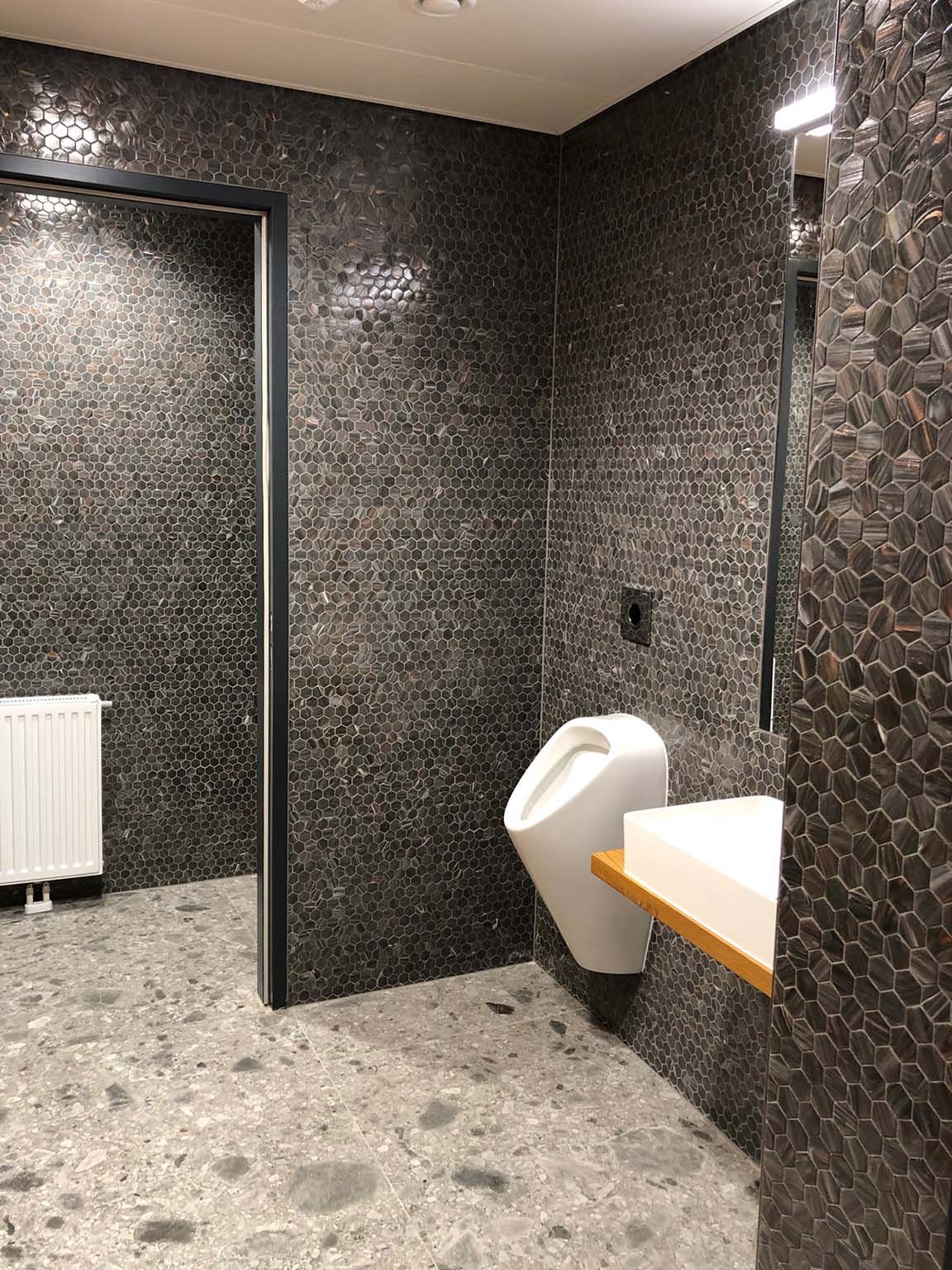 Ein Badezimmer mit Fliesen in Mosaikoptik an den Wänden, mit Urinal, Waschbecken und Spiegel.