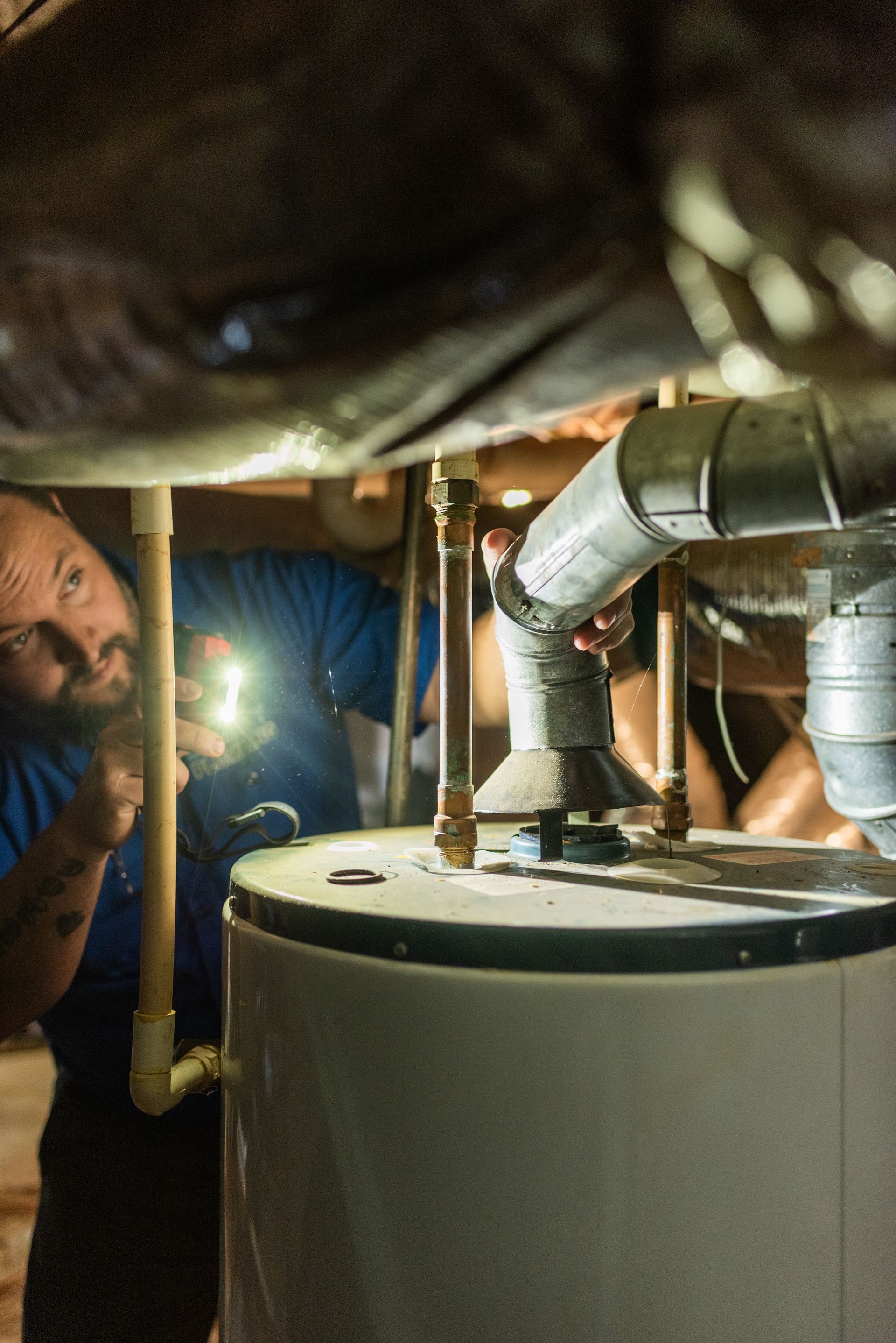 A man is looking at a water heater in a basement with a flashlight.