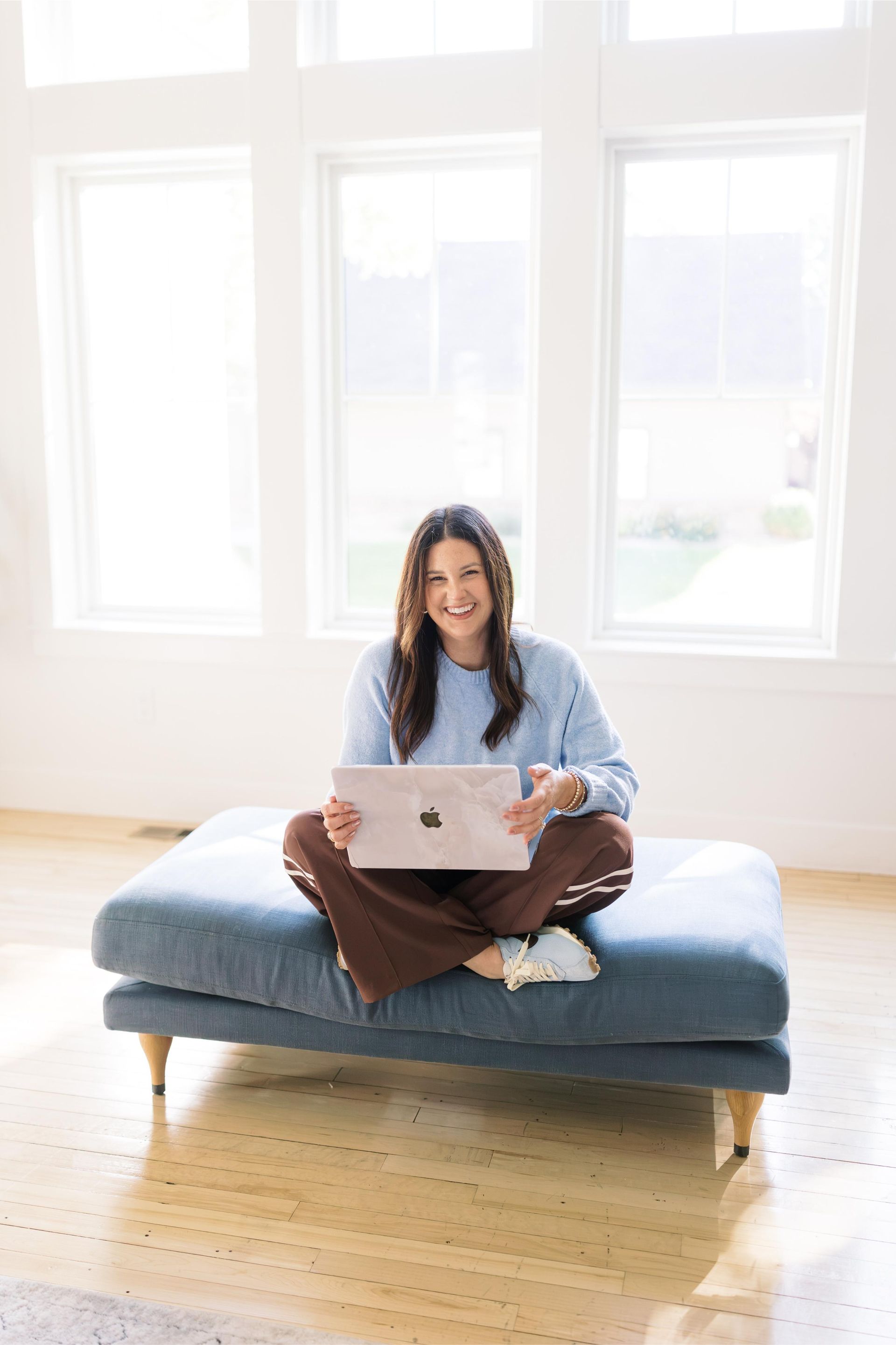 Woman sitting on a blue bench using a laptop in a bright room with large windows.