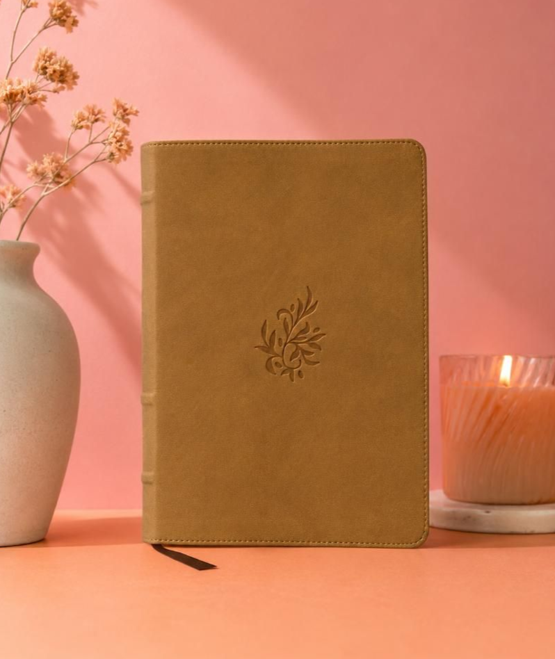 Tan notebook with leaf embossing on a pink tabletop beside a vase and lit candle