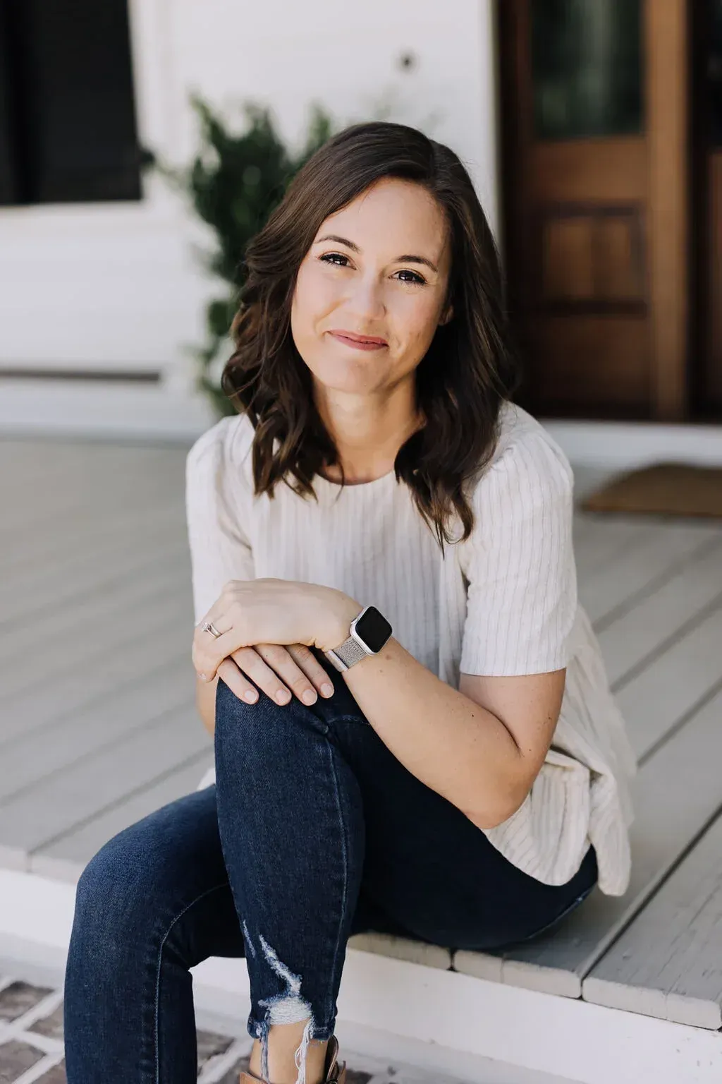 Woman sitting on porch steps, smiling in a cream top and jeans.