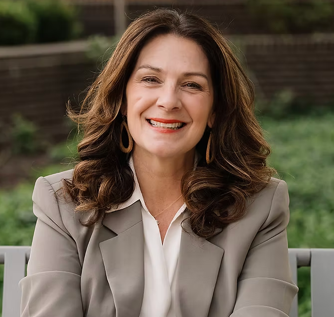 Smiling woman in a beige blazer seated outdoors against a blurred garden backdrop