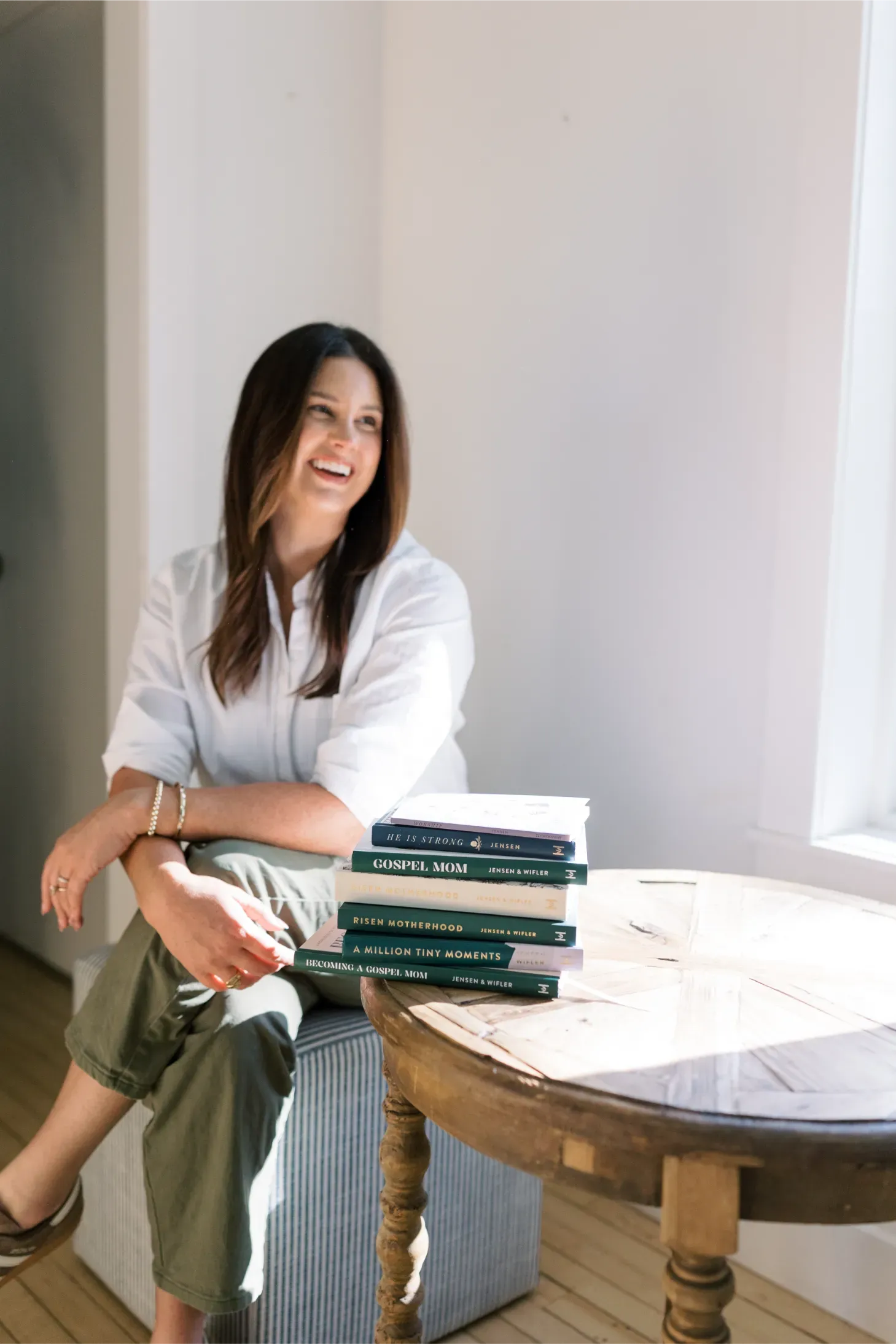 Woman sitting by a bright window beside a stack of books on a wooden table