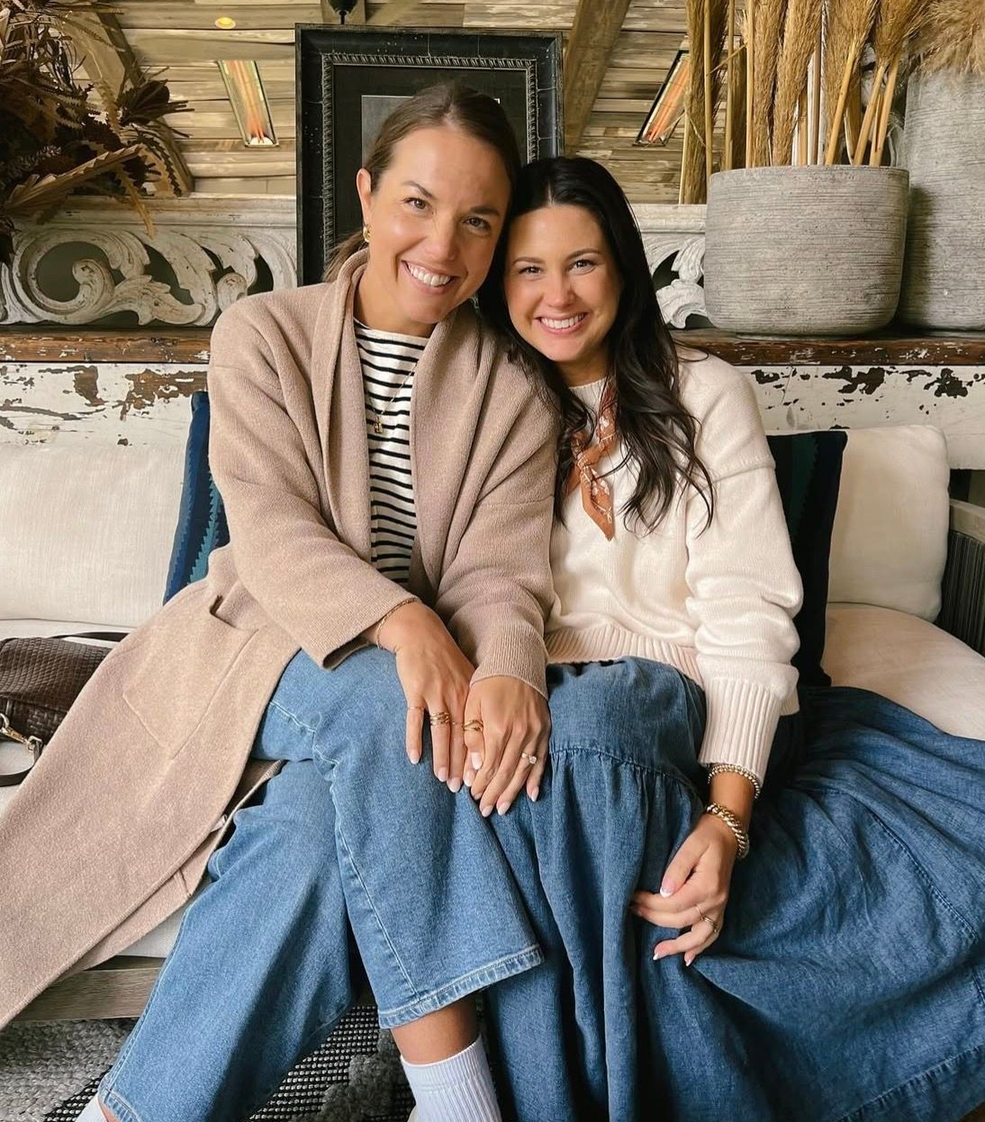 Two women smiling on a couch in a cozy rustic room, wearing beige and denim outfits.