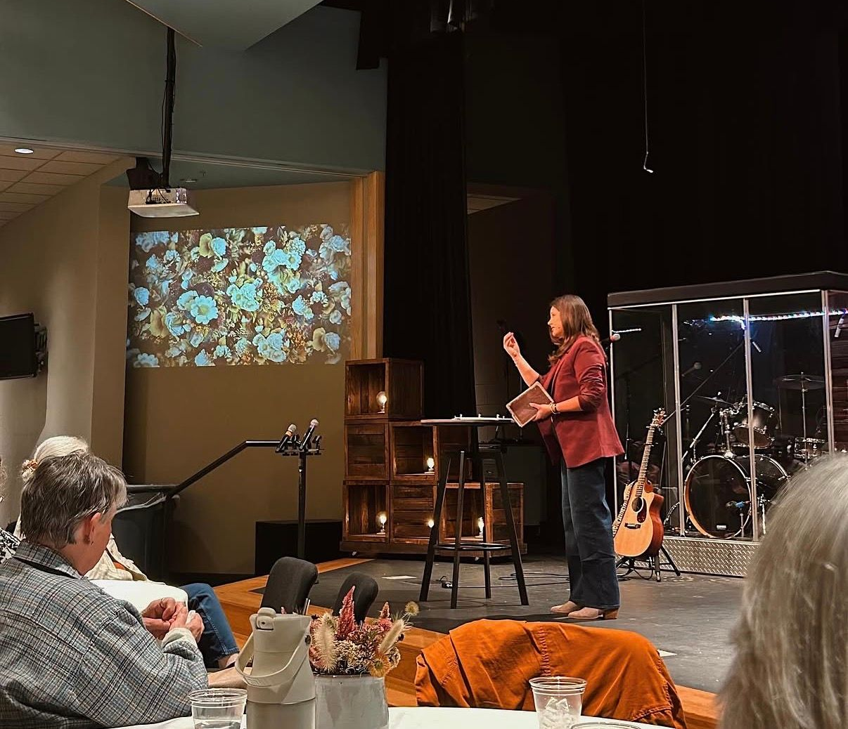 Man speaking on a church stage beside a drum booth, with a projected floral image behind him and audience in front