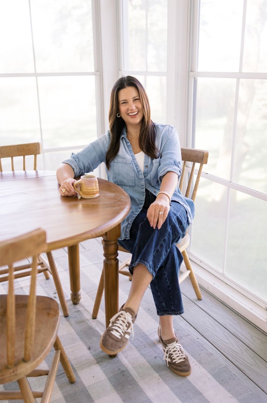 Woman smiling at a round wooden table in a bright sunlit room, holding a drink and wearing jeans and sneakers