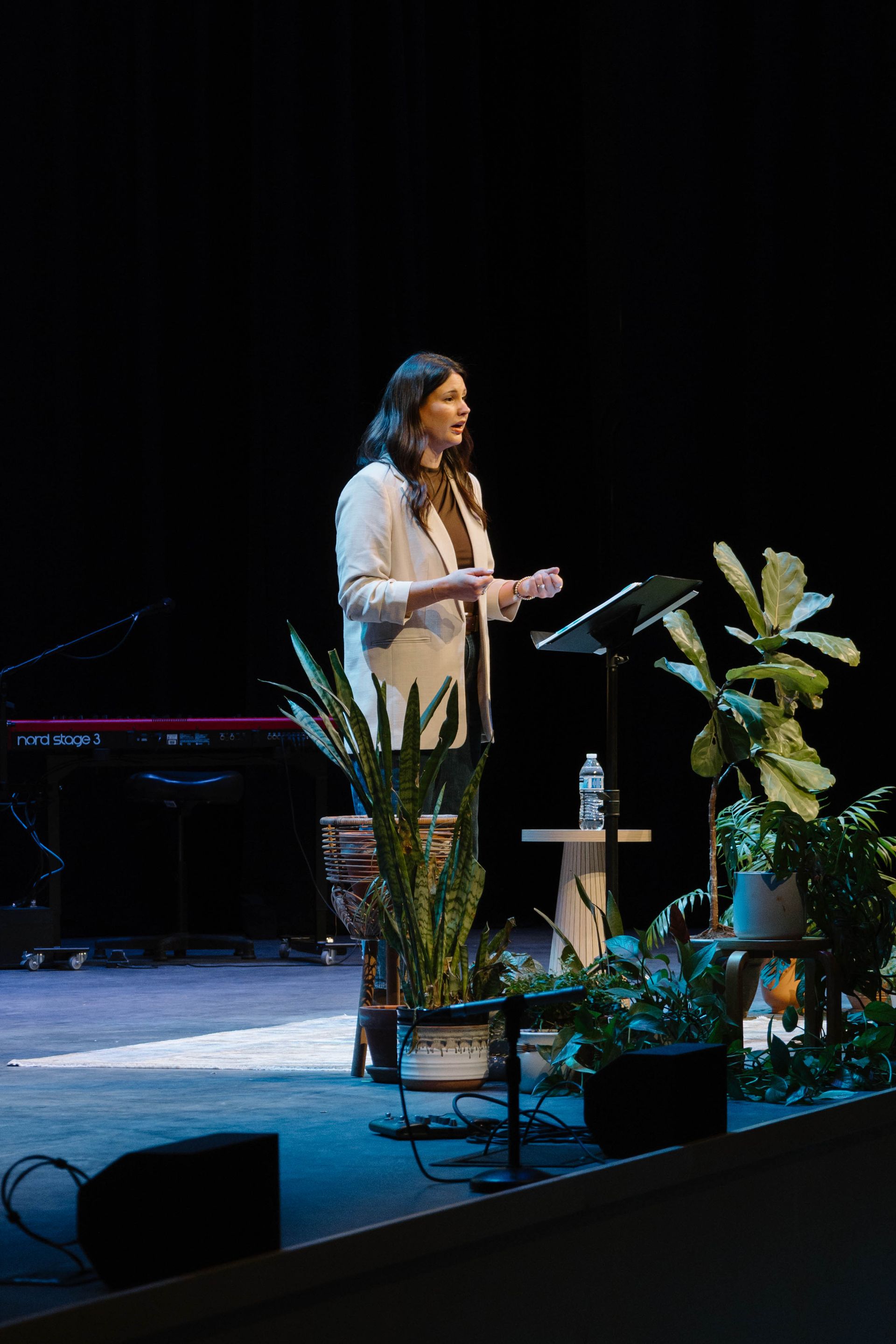 Woman speaking on a stage beside potted plants, holding notes under blue lighting