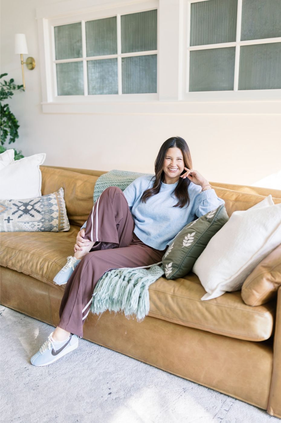 Woman sitting on a tan couch in a bright living room, smiling with pillows and a throw blanket around her.