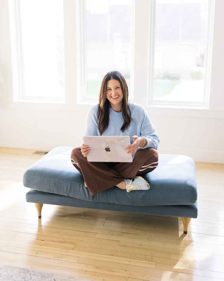 Woman sitting on a blue bench using a laptop in a bright room with large windows.