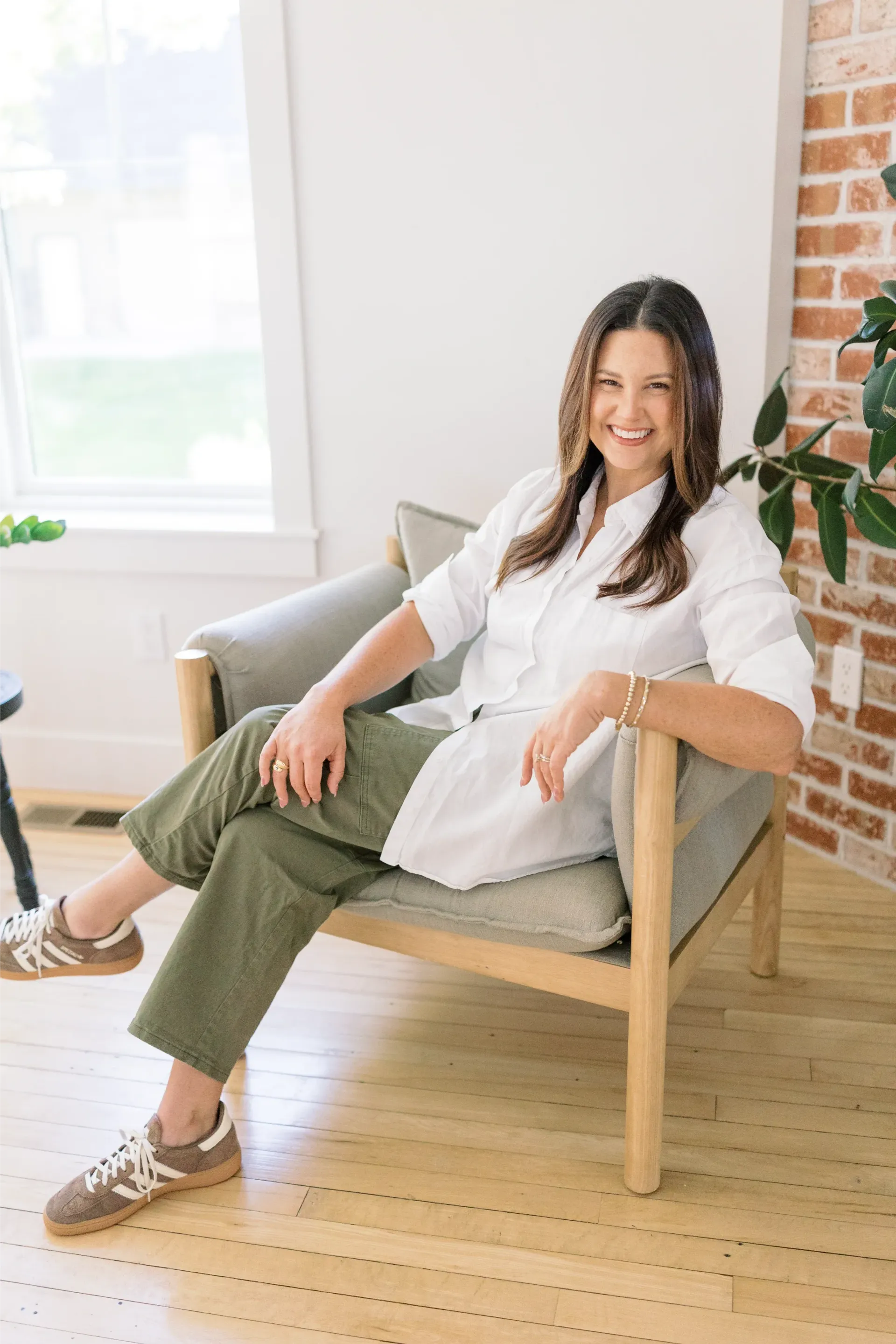 Woman sitting in a light wood chair in a bright living room, smiling and relaxing by a brick wall.