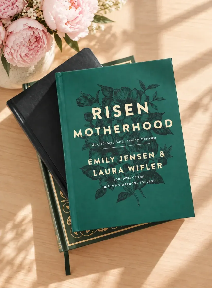 Two stacked books on a sunlit table, the top a green book titled “Risen Motherhood” with pink flowers nearby