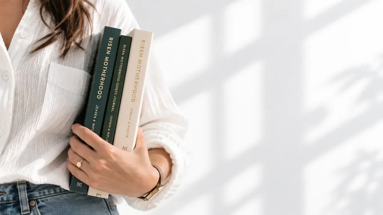 Person in white shirt and jeans holding three books by a sunlit window