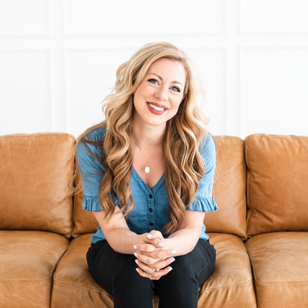 Smiling woman in a blue shirt seated on a tan couch against a white wall