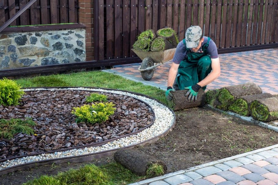 Gardener kneeling to plant shrubs in a landscaped yard with a curved stone border.