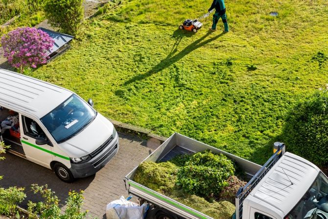 Overhead view of a gardener mowing a bright green lawn beside white vans and flower beds