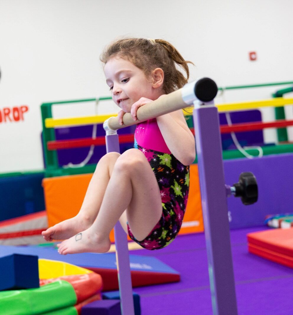 Young girl in gymnastics leotard on low uneven bars, legs bent, smiling. Gym setting.
