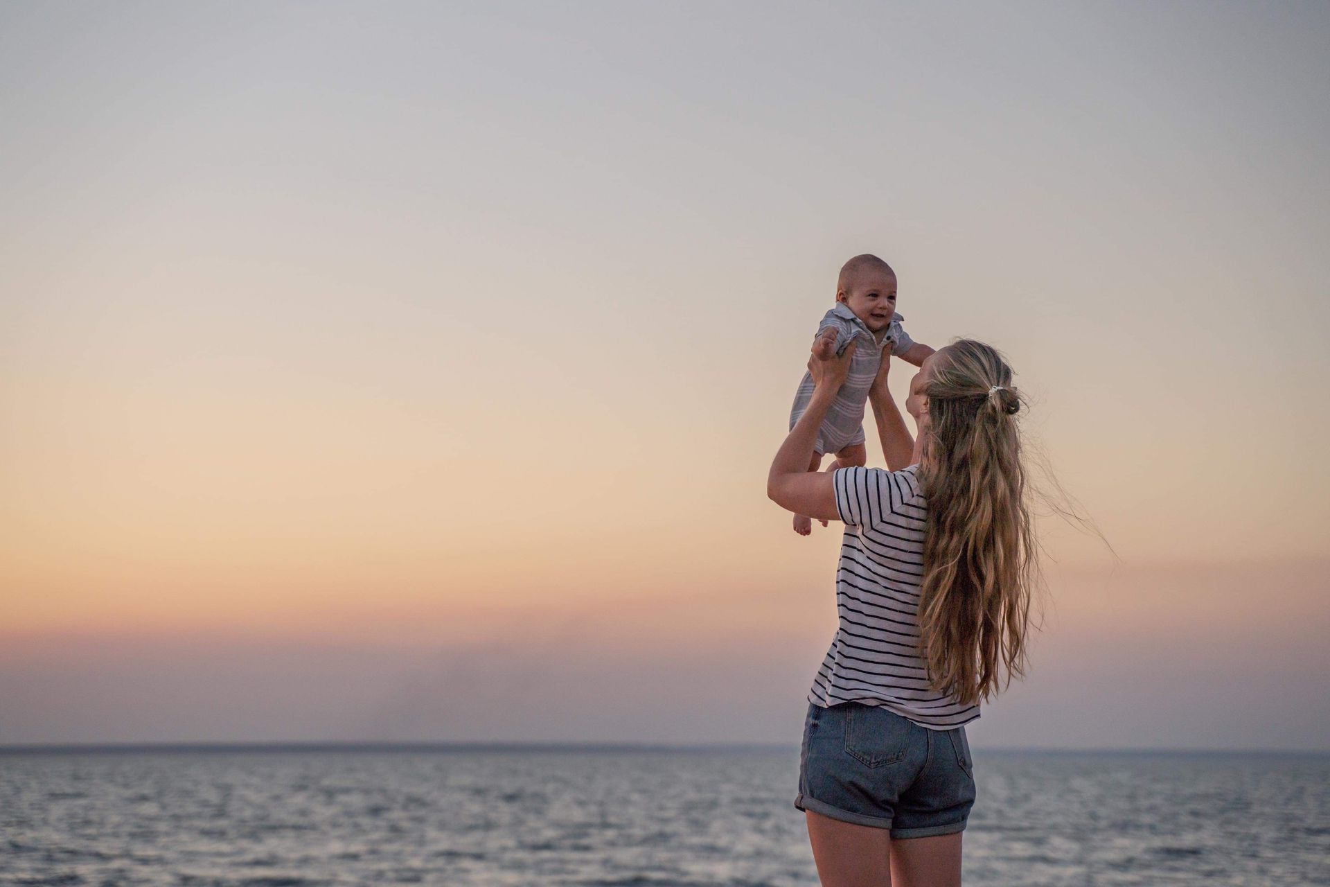 Adult lifting a baby at the beach during sunset, with calm water and a colorful sky.