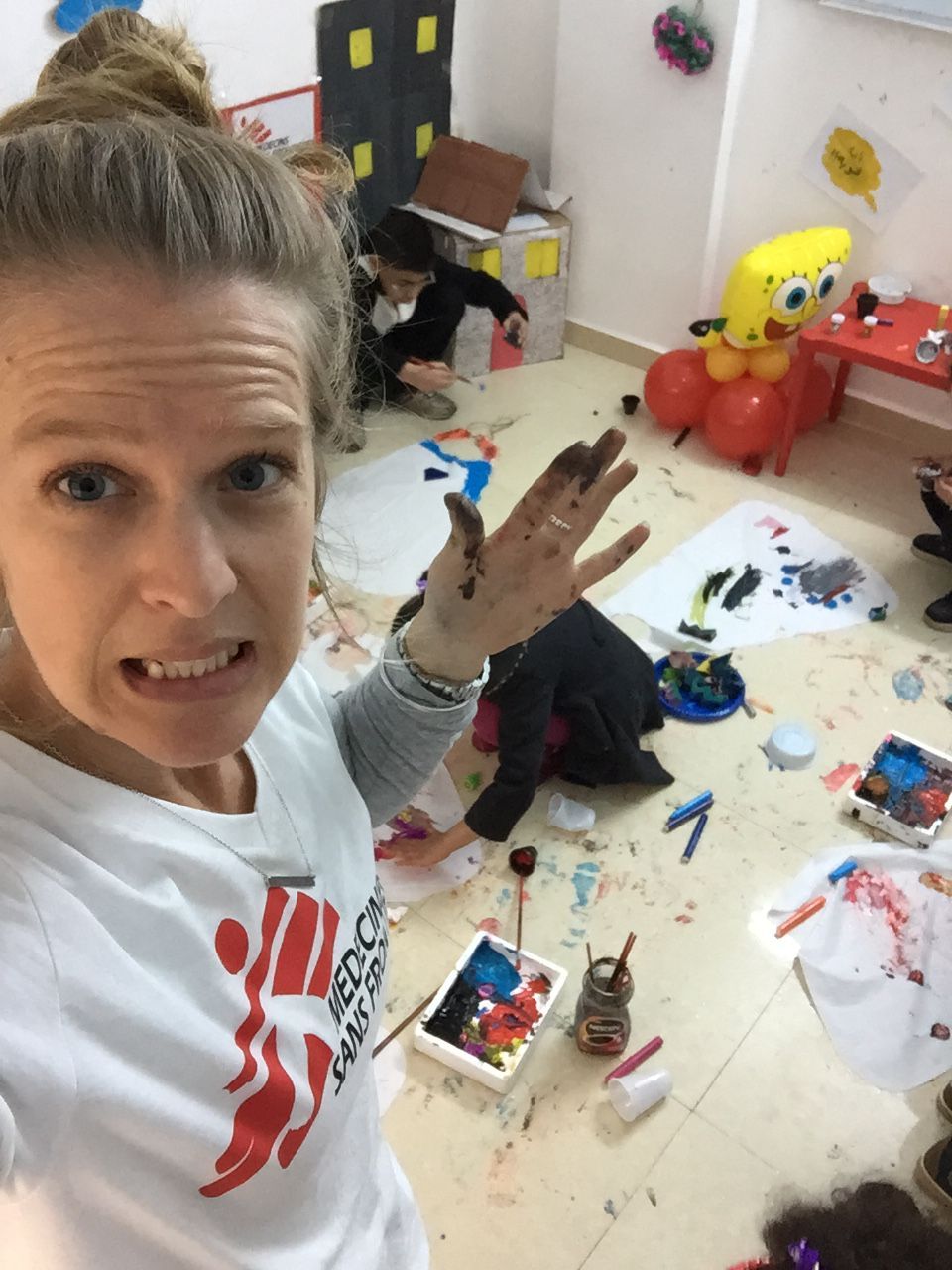 Adult in a messy craft room taking a selfie while children play on the floor with art supplies.