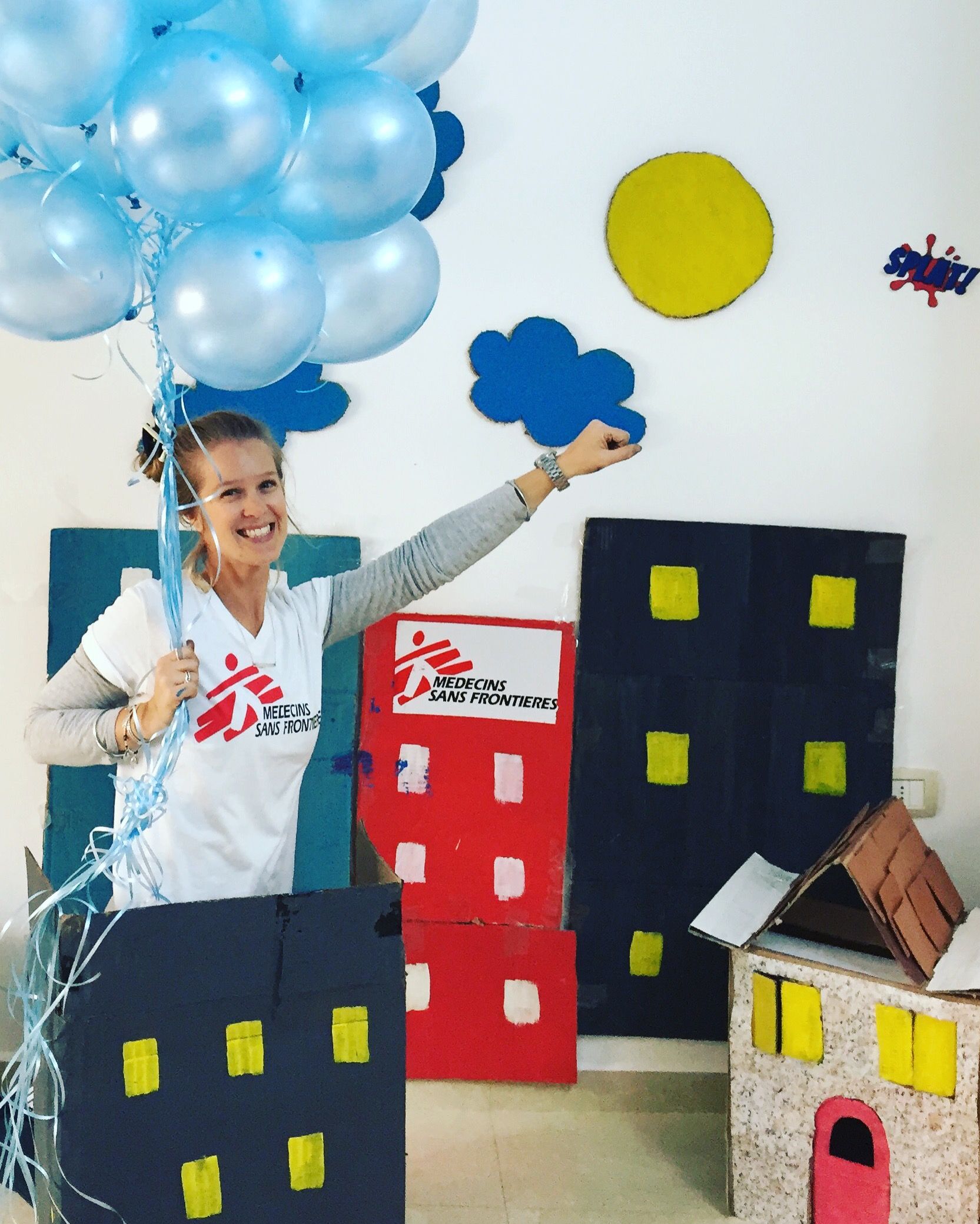 Person in a cardboard city scene holding blue balloons and a cloud prop against a white wall.