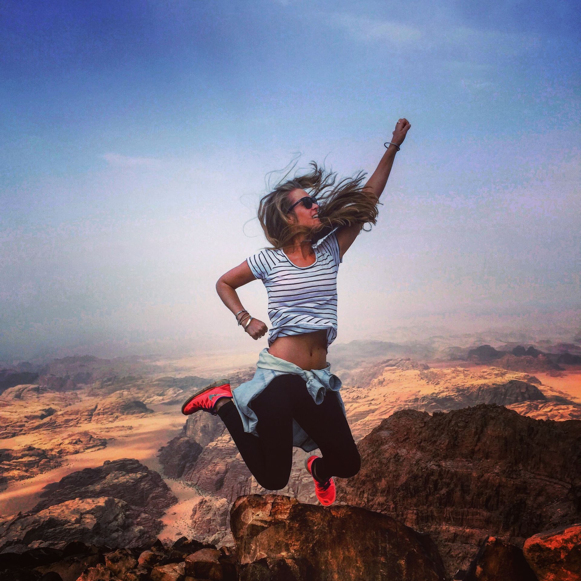 Person mid-jump on rocky desert cliff with one arm raised, wearing striped shirt and red shoes