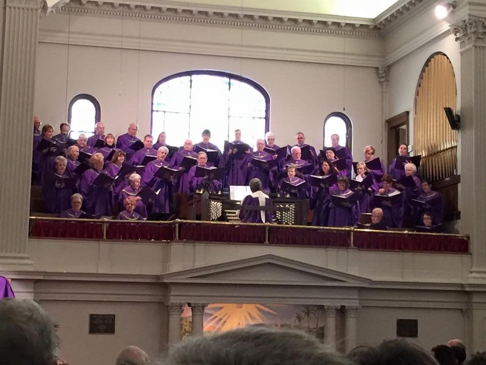 Choir in purple robes singing in a large hall with arched windows.