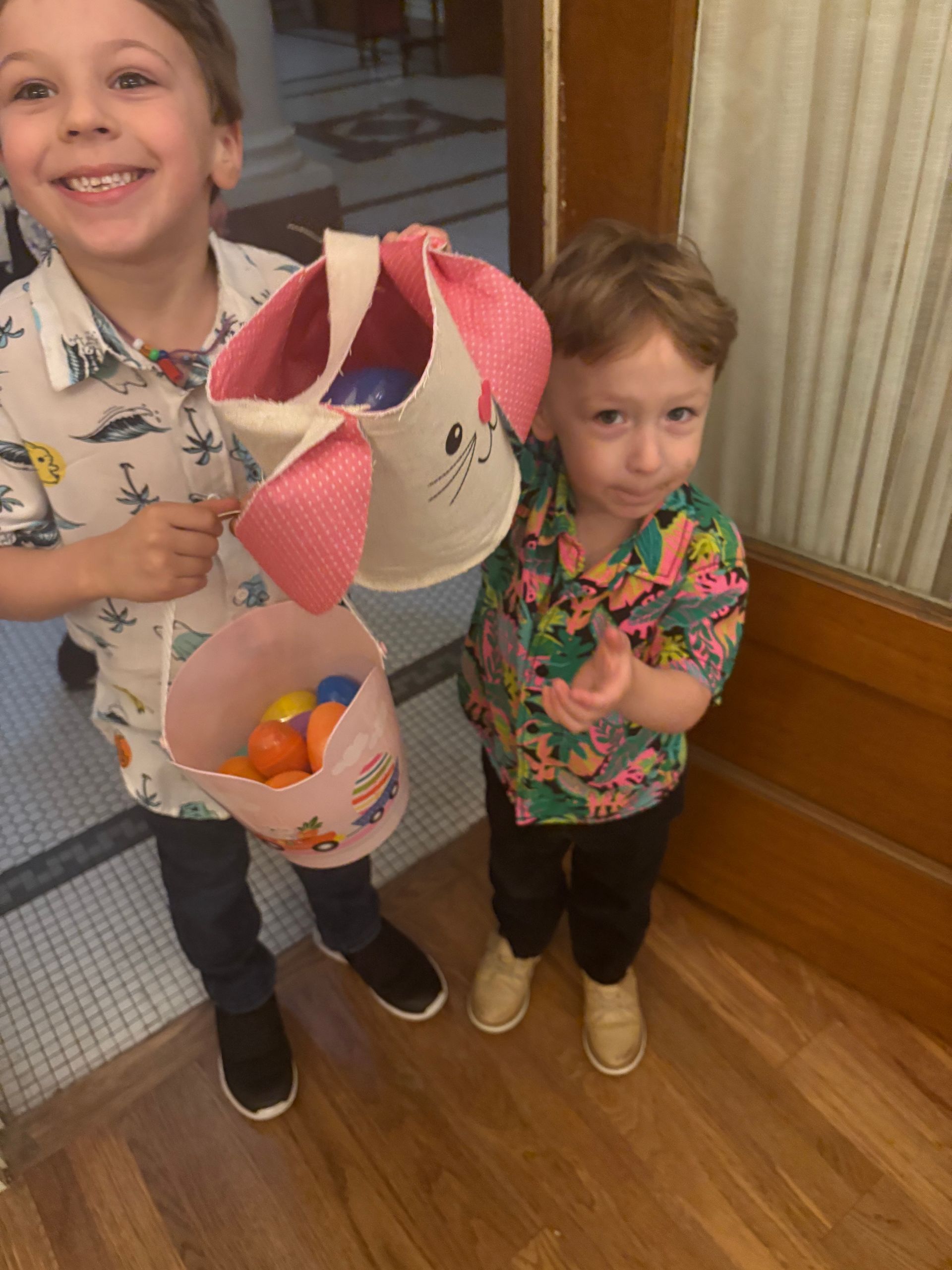 Two young children with Easter baskets, one smiling, holding a bunny-eared basket, the other looking unsure.