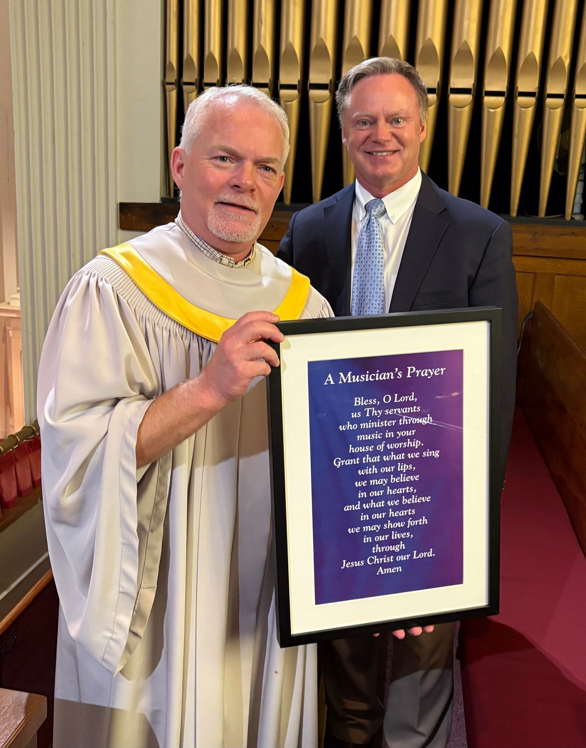 Two men in a church, one in vestments holding a framed poem, smiling. The other man is in a suit.