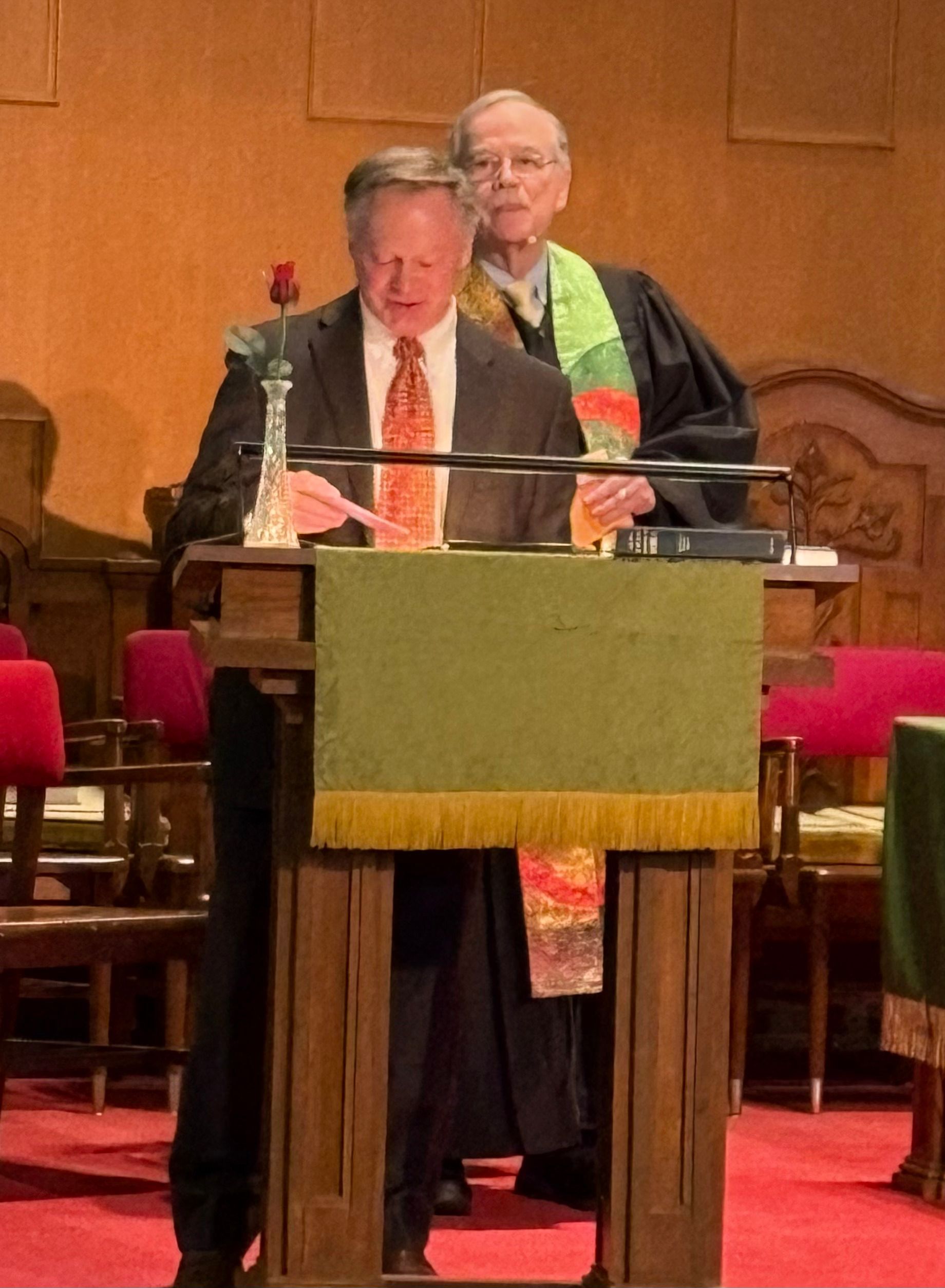 Man speaking at a lectern with another man behind him in a church. Red rose in a vase.