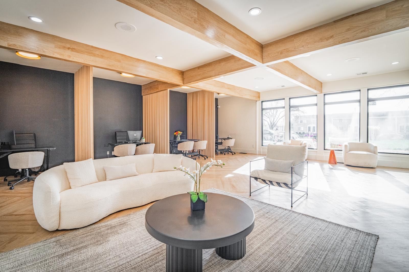 Modern open office lounge with white sofas, desks, and large windows under wood ceiling beams