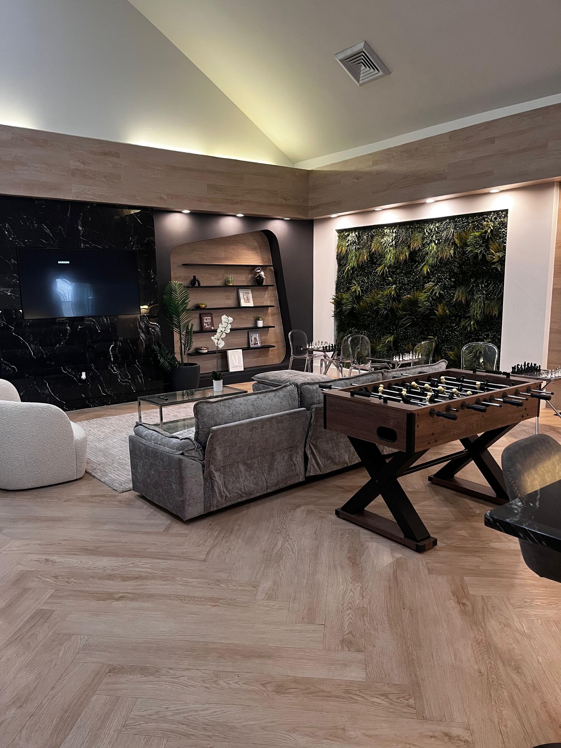 Modern living room with gray sofas, a pool table, wall shelves, and a green plant wall.