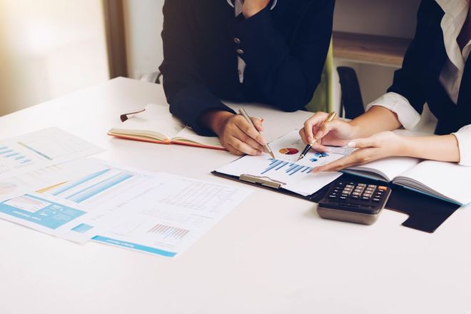 Two people in business attire review financial charts and data at a desk with a calculator.