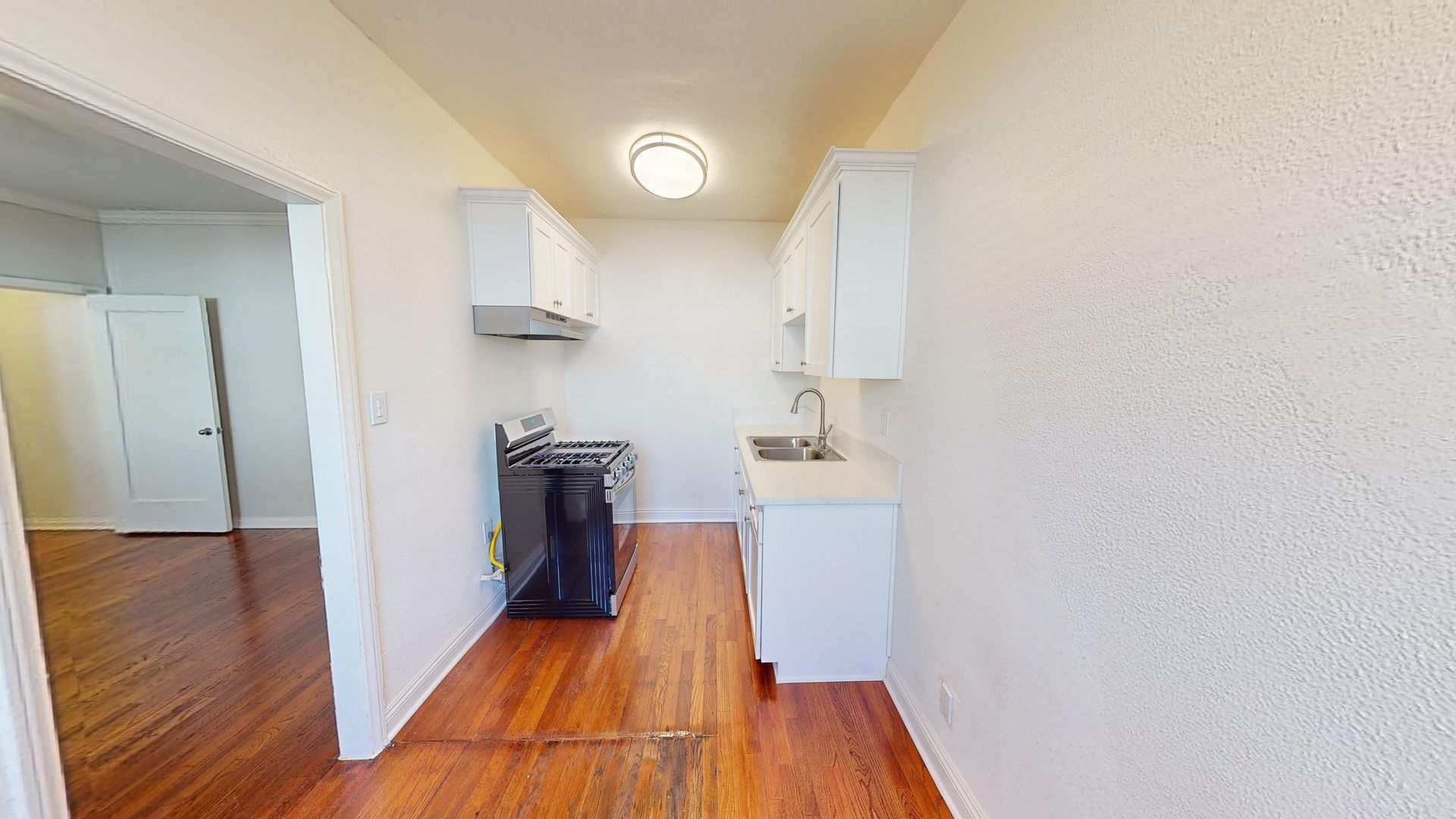 Small kitchen with white cabinets, stove, sink, and hardwood floors. Doorway to another room.