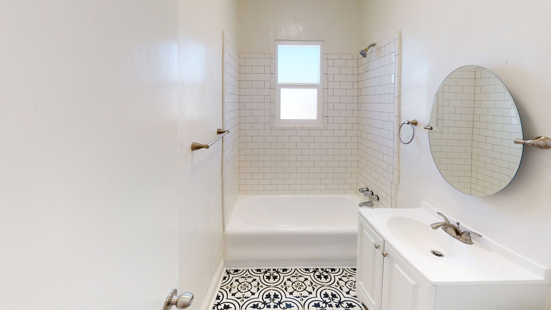 White bathroom with patterned floor, bathtub, sink, and round mirror. Window above bathtub.